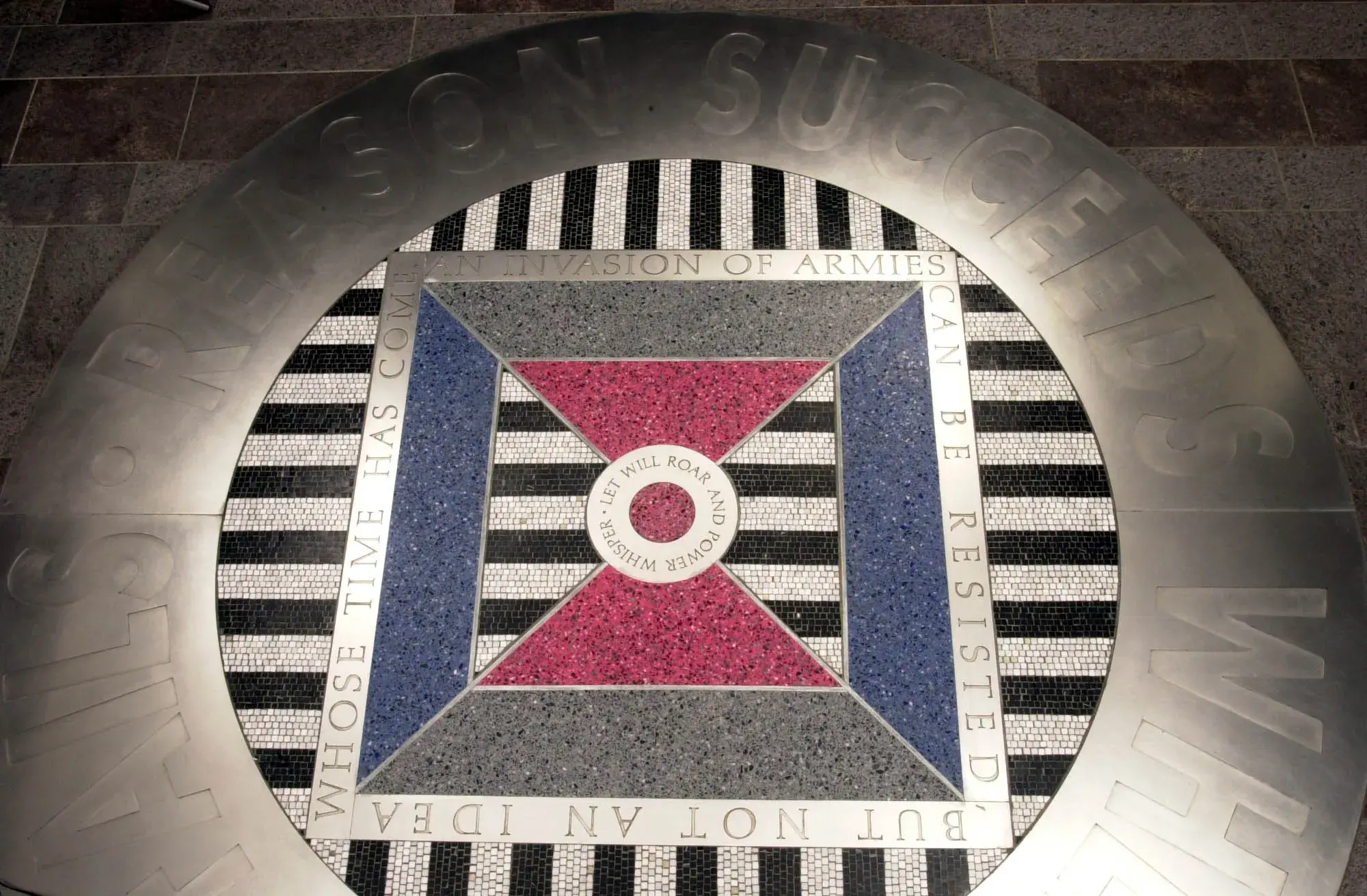 Circular floor installation with thick metal band around the edge and a black and white and red, blue, and gray mosaic and terrazzo pattern in the center. The words "Let will roar and power worship" are visible in the center.