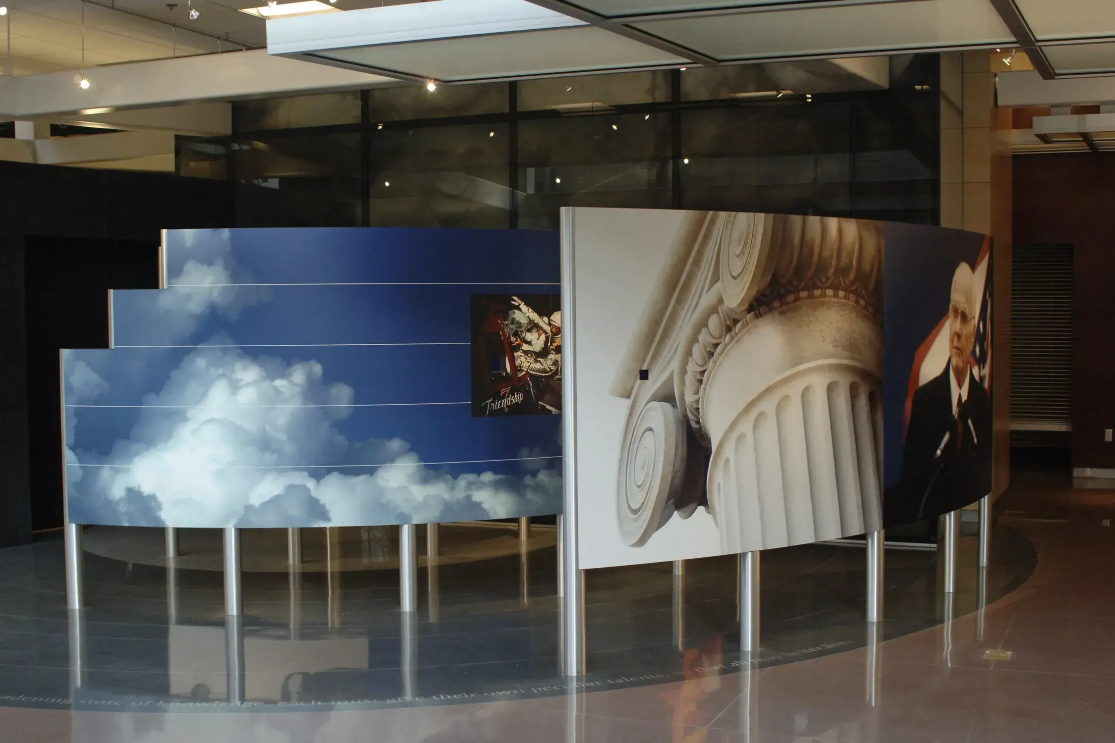 Images of clouds, Friendship 7 space capsule, an Ionic capital, and an older  man wearing glasses and a suit in front of a red, white and blue flag are printed on 2 curved panels held up on metal legs. The shiny floor has a large circle on it.