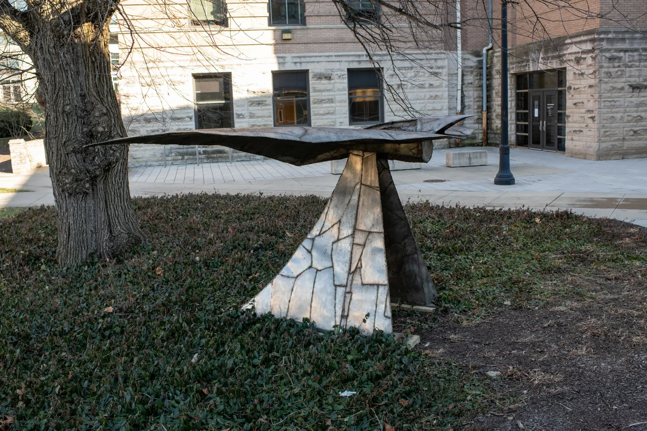 Metal sculpture sits in a landscape bed next to a tree near a stone building. The sculpture has a horizontal component on top of a sloping vertical base.