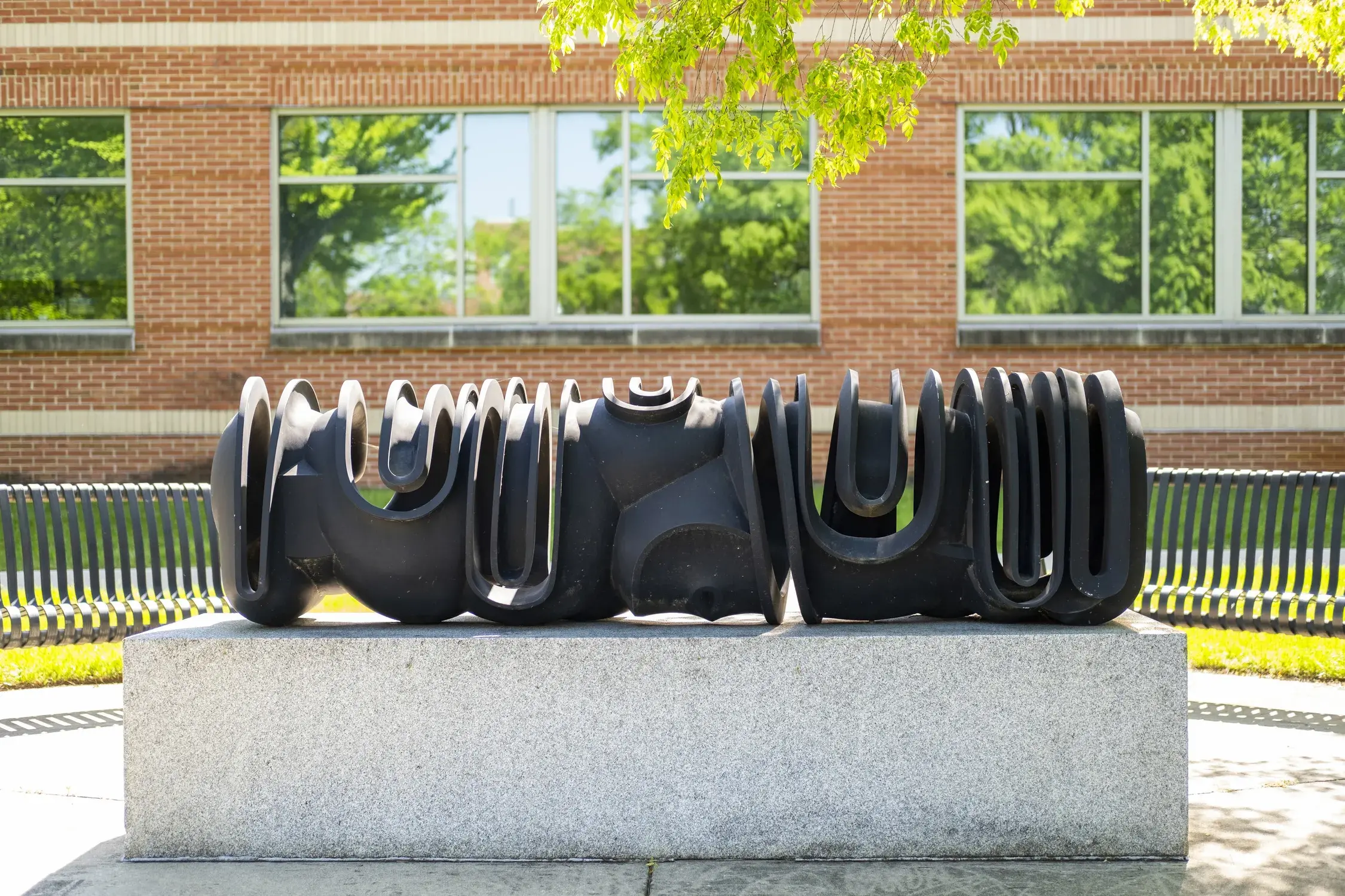 Dark metal sculpture of curvilinear forms sits on a gray stone base in front of a brick building