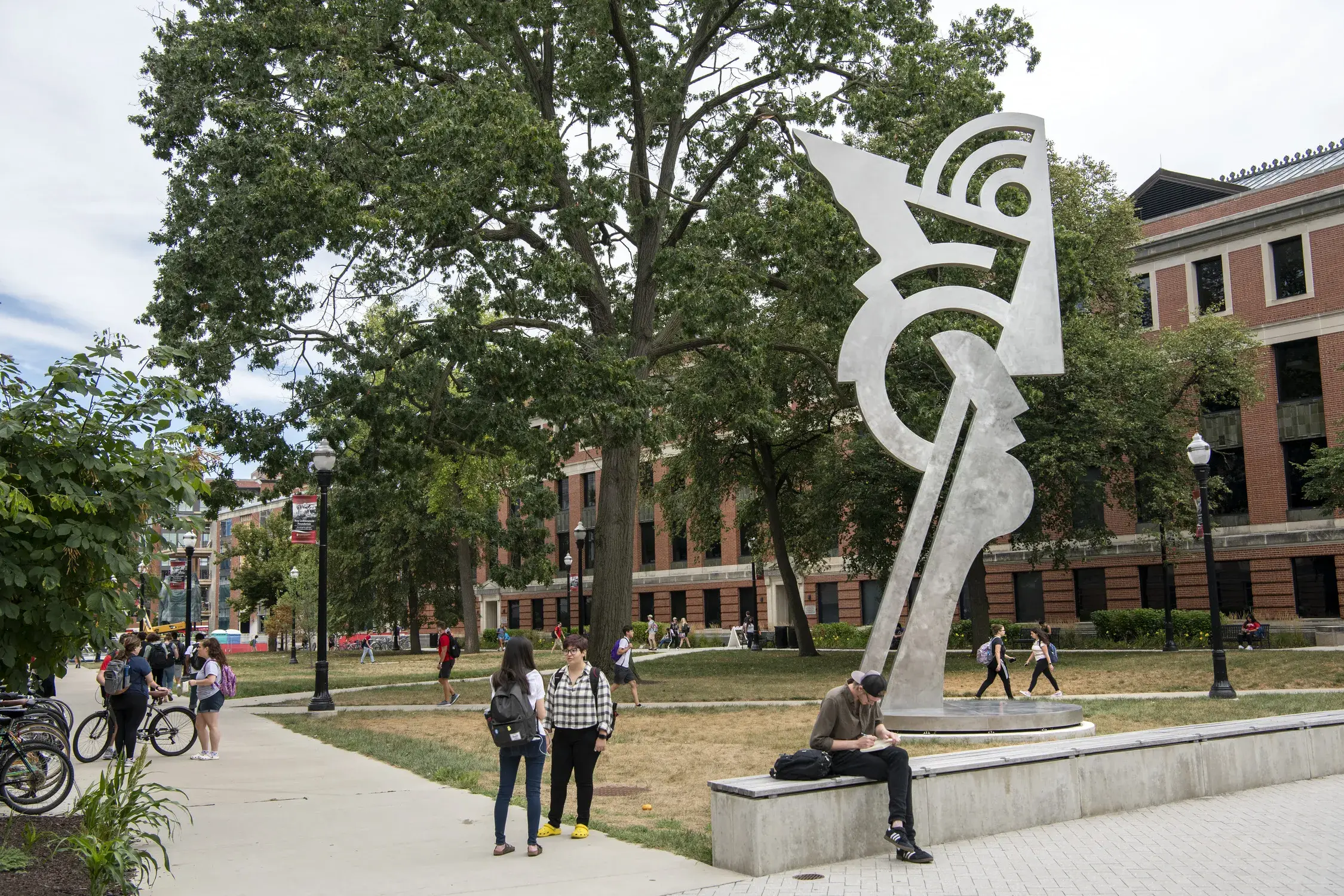 Large silver colored sculpture in the shape of an abstract head on a long neck. There are trees and several-story-high brick buildings in the background and several people walking and sitting near it.