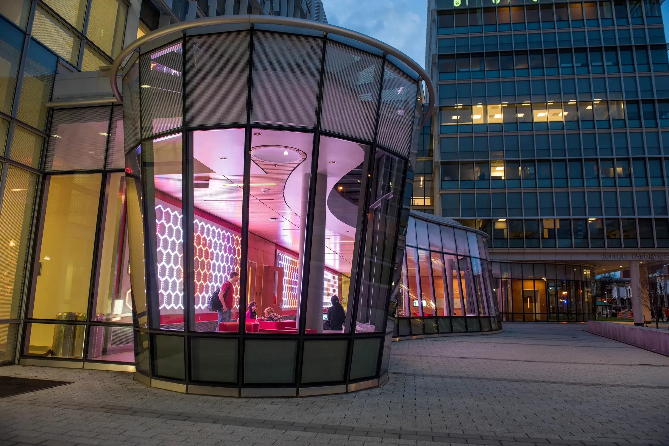 View from outside a curved glass wall into a lobby space with three walls of a light installation consisting of small bulbs in a honeycomb pattern. The lights have different patterns of colors and reflect onto the ceiling. People gather in a circular area in the foreground.