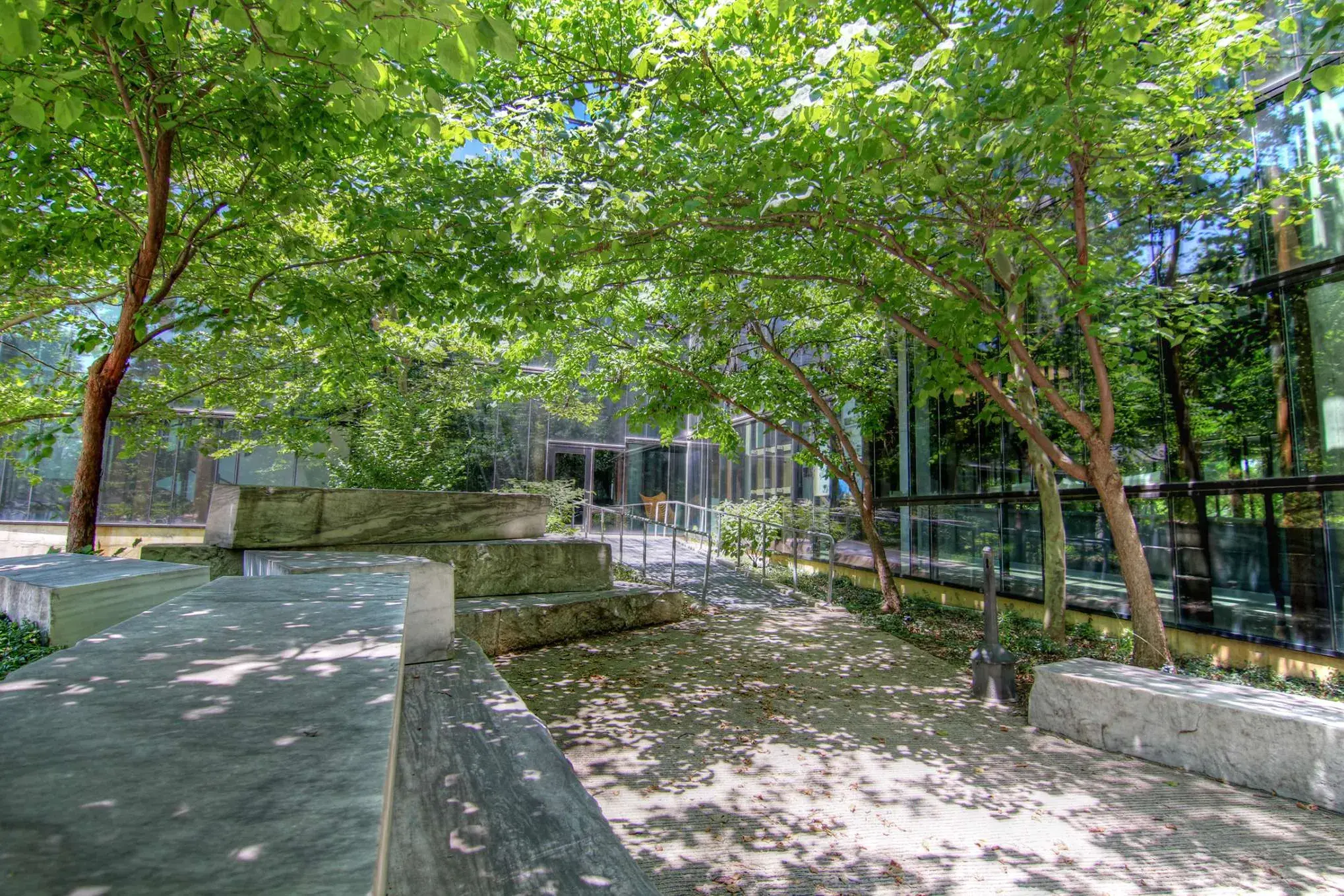 Slabs of white and gray marble which sit alongside a walkway in a courtyard with trees, next to a glass building wall 