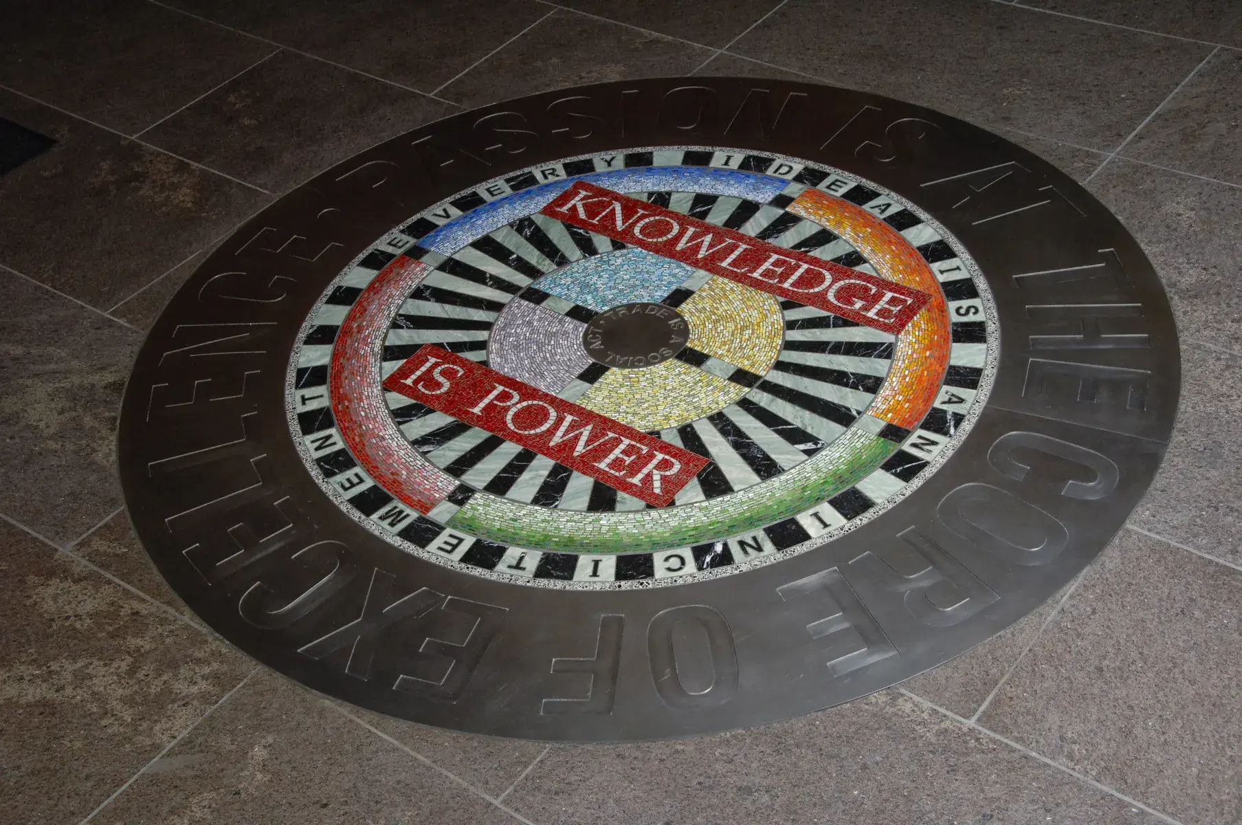 Circular floor installation with thick metal band around the edge and a black and white and multicolored mosaic pattern in the center. The words "knowledge is power" are visible.