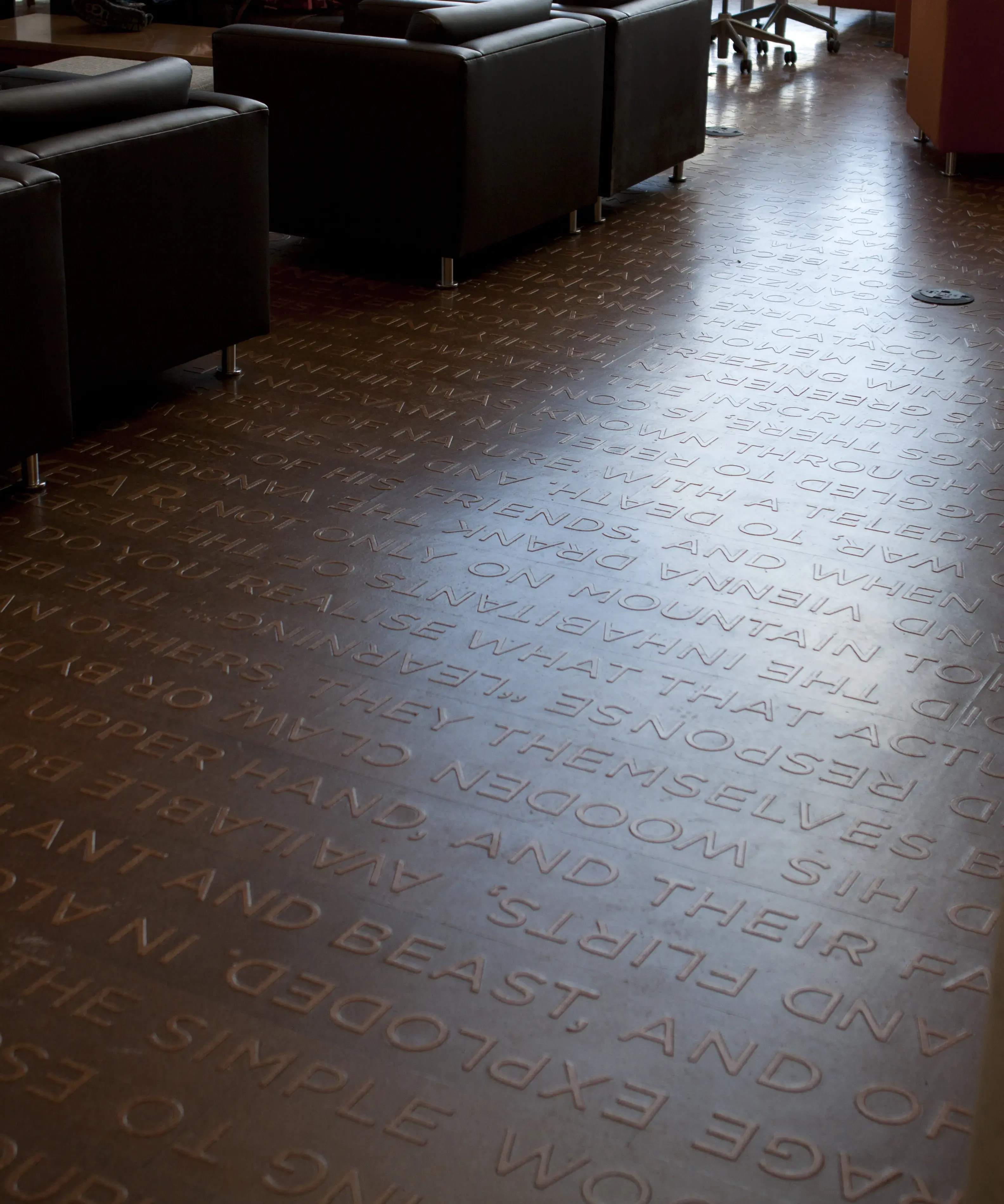 Medium brown floor with lines of lighter brown words slightly raised up from the surface. There are chairs and other furniture in the background with people using some of them.