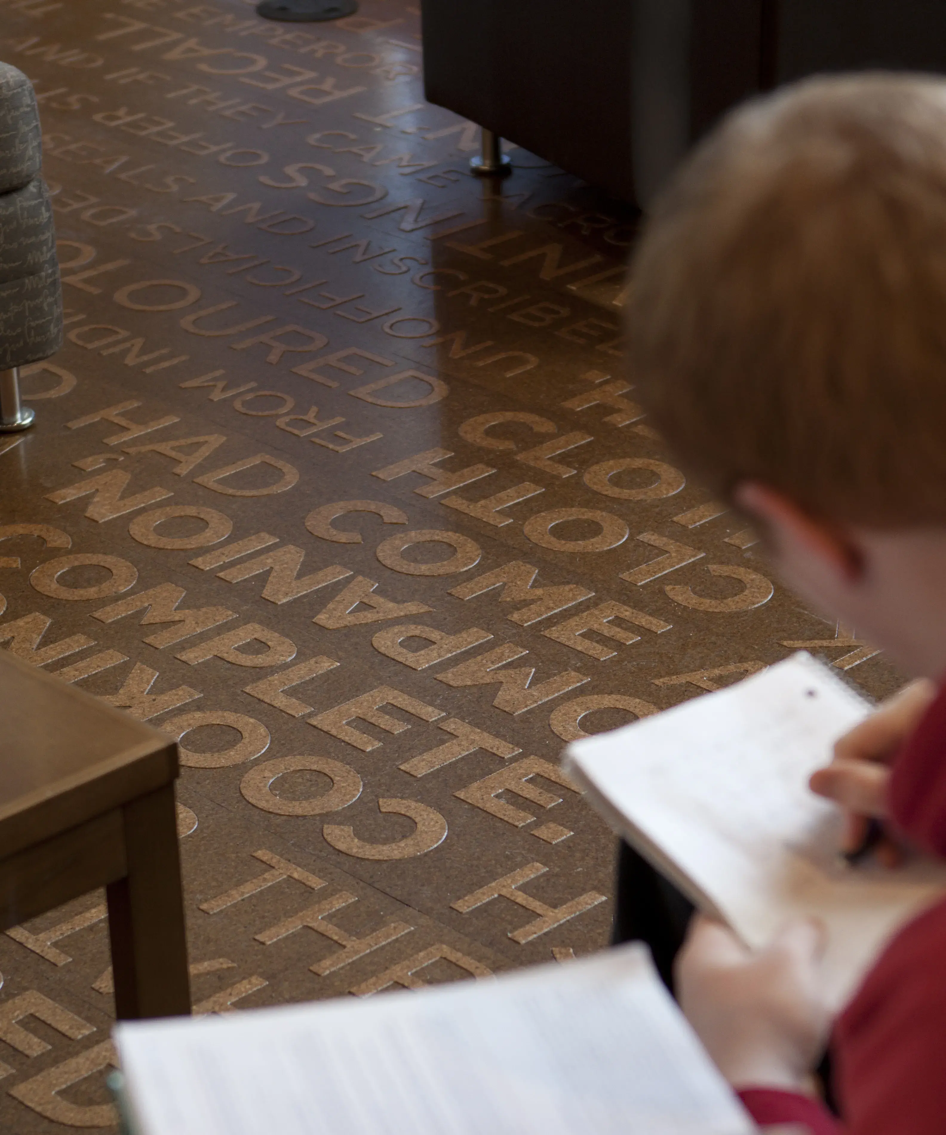 A person is writing on a notebook over a medium brown floor that has lines of slightly raised lighter brown words on it.