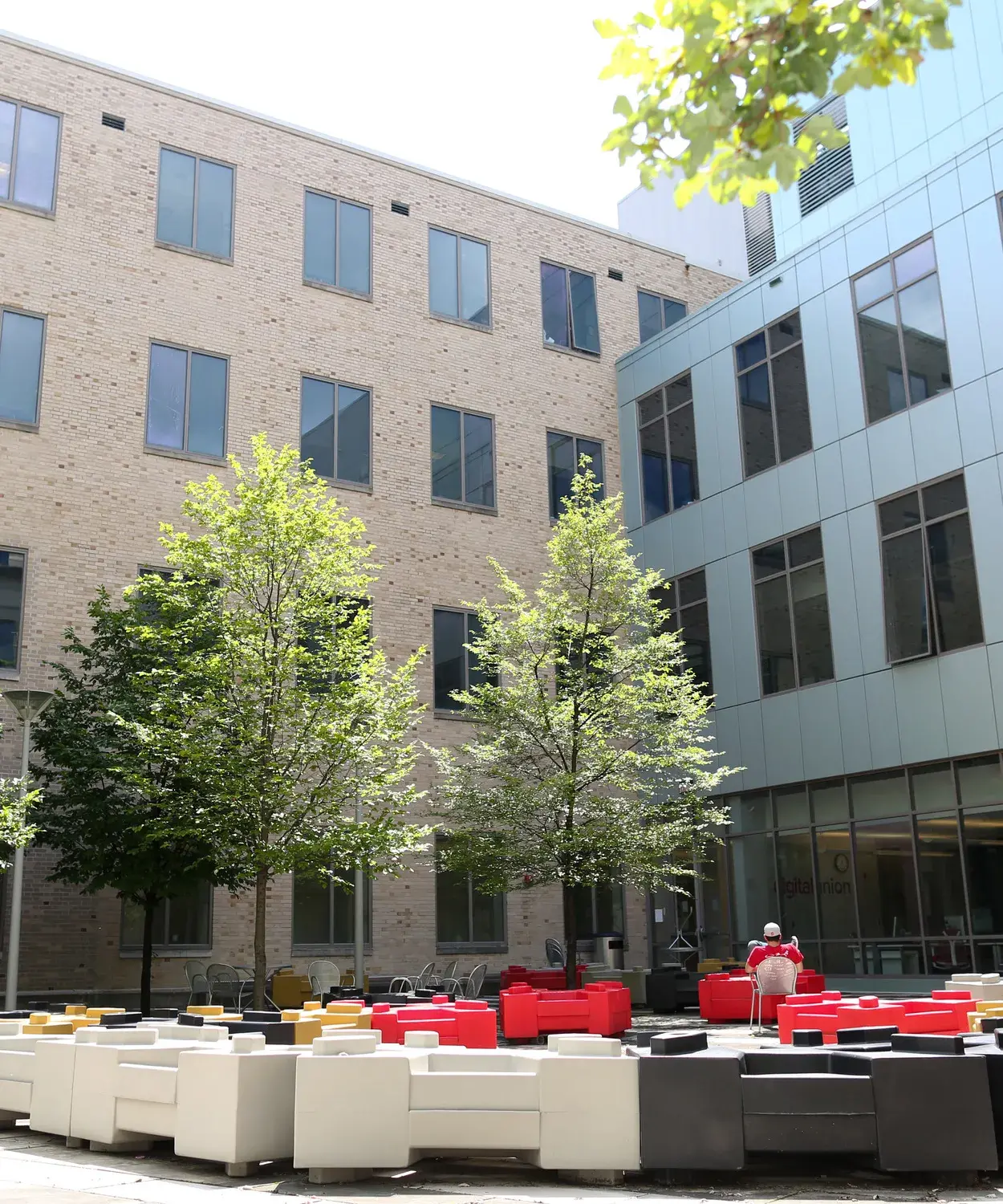 Red, yellow gold, gray, and black  objects sit on a courtyard in front of a building with one metal-clad wall and one brick wall. There are trees and chairs as well.