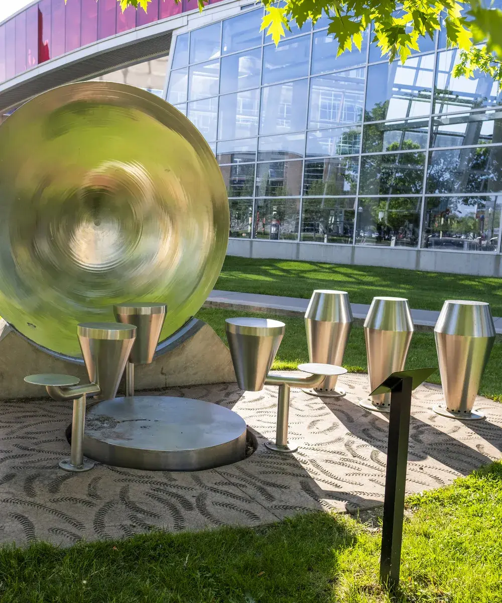 Multiple silver metal drums of various types sit on a textured area in the grass. There is a glass wall of a buildings behind it.