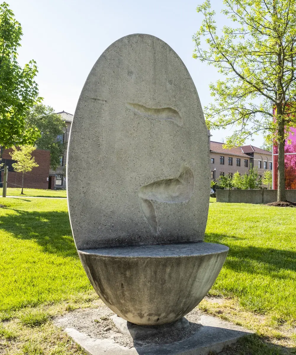 Oval concrete sculptural bench with an impression of the side of a person's arms and legs in a squatting position on the vertical back portion