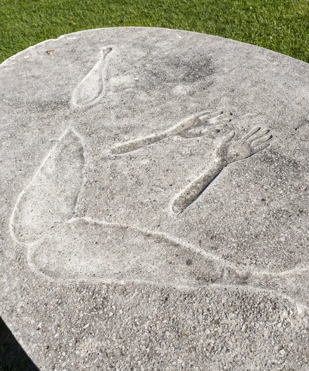 Round concrete sculptural bench with an impression of a person's body bending over on the vertical back portion, with another round concrete piece without a back in the foreground 