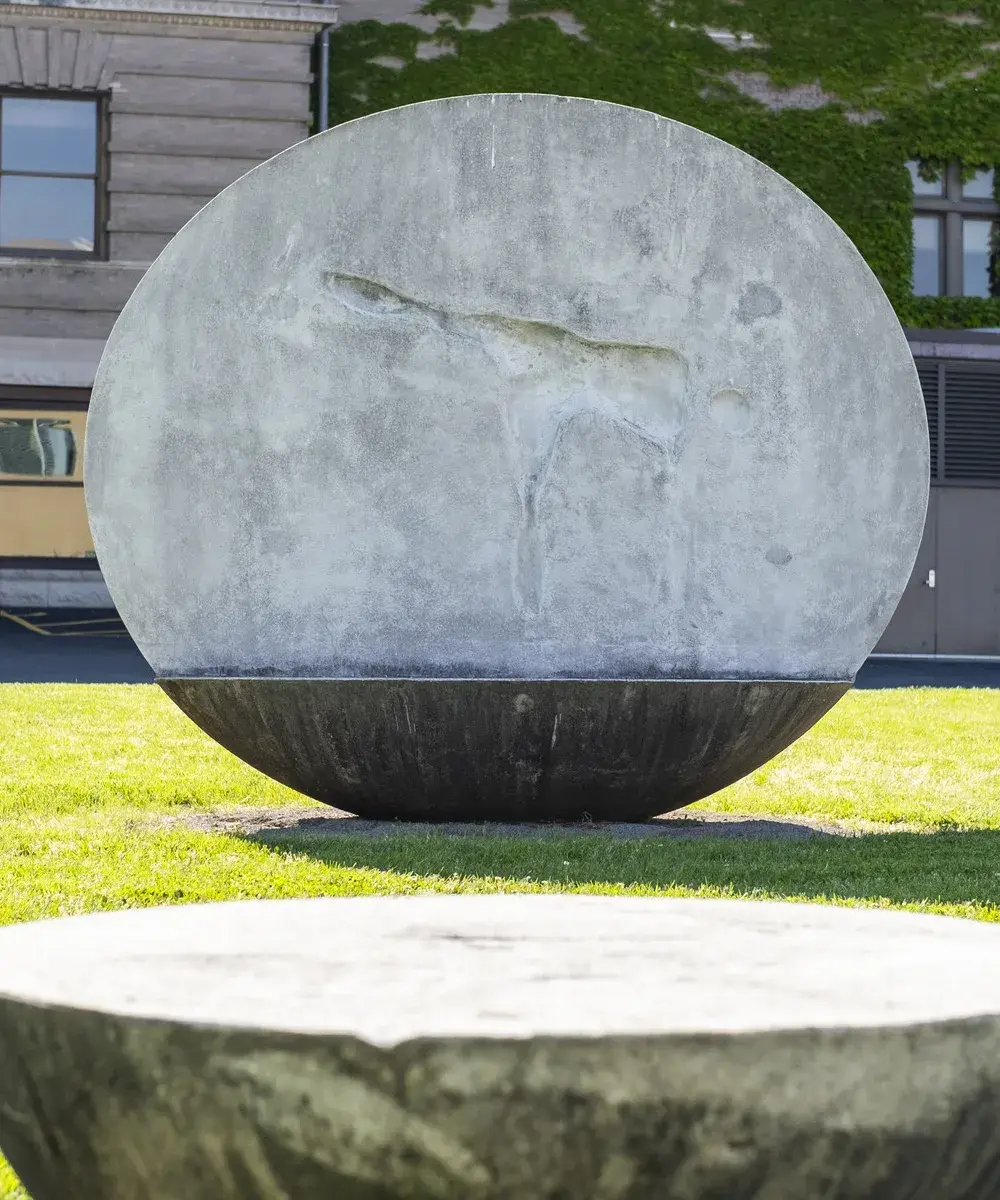 Round concrete sculptural bench with an impression of a person's body bending over on the vertical back portion, with another round concrete piece without a back in the foreground