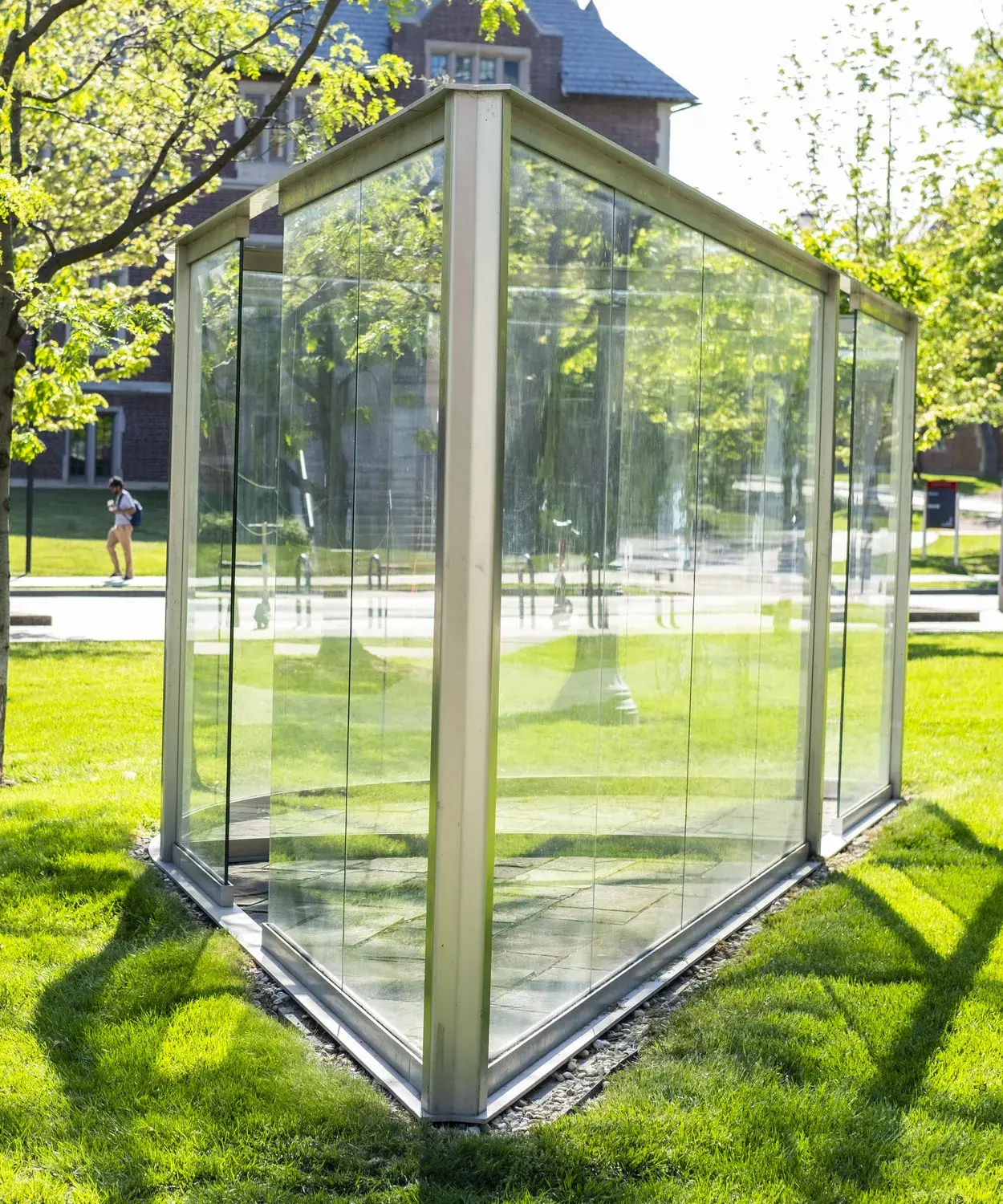 Large glass panes within a metal frame come to a point on one side and extend back, with one side curving. The sculpture sits in a grassy area.