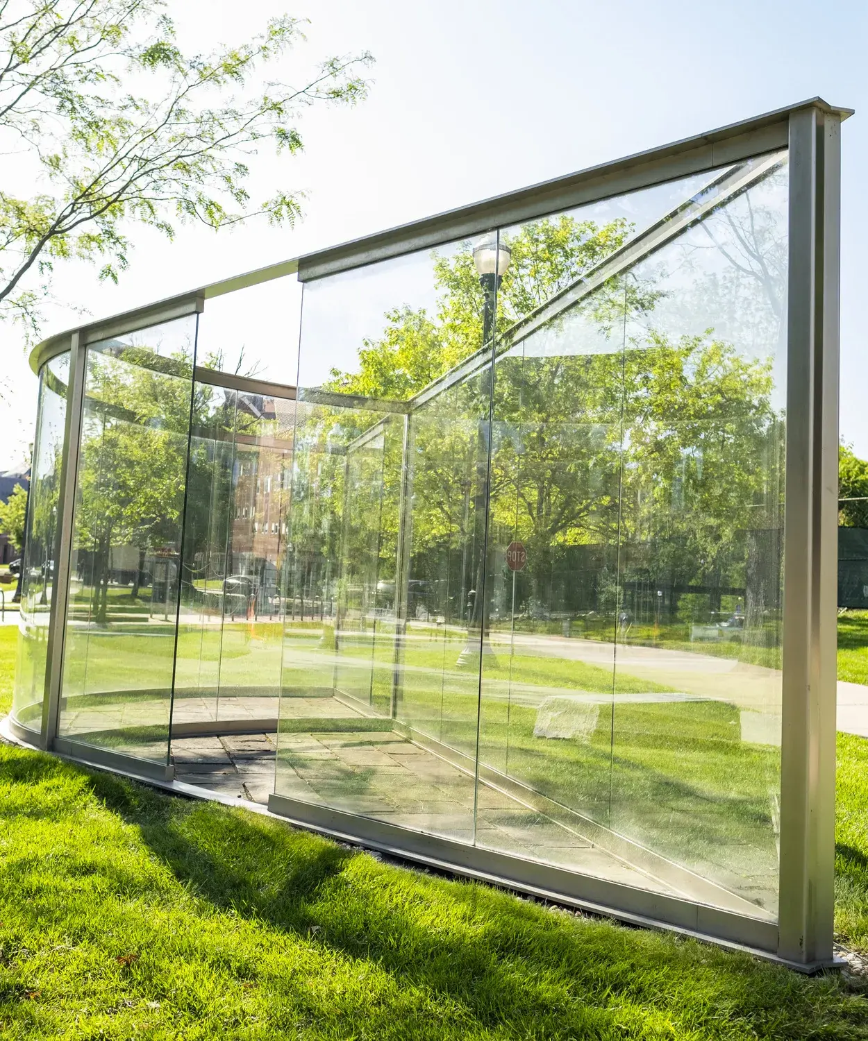 Large glass panes within a metal frame come to a point on one side and extend back, with one side curving. The sculpture sits in a grassy area.