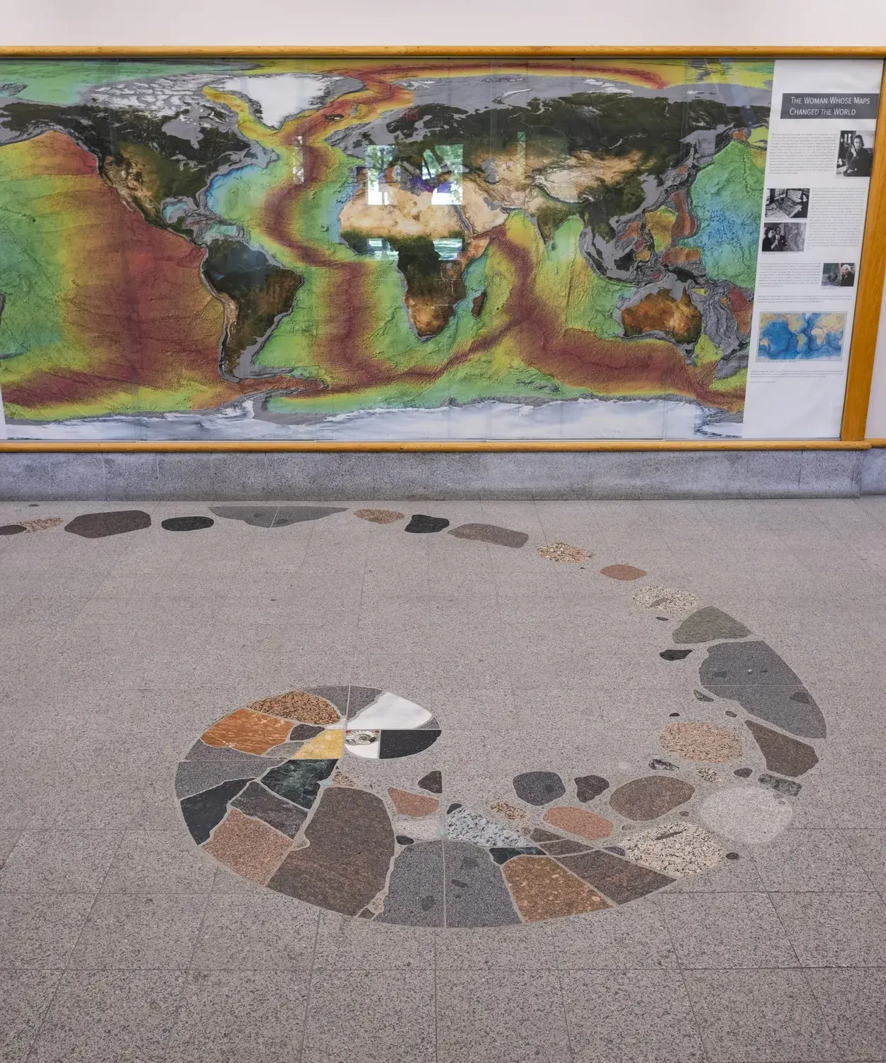 A floor design with a spiral shape made of different types and colors of rocks. There is a map on the wall in the background.