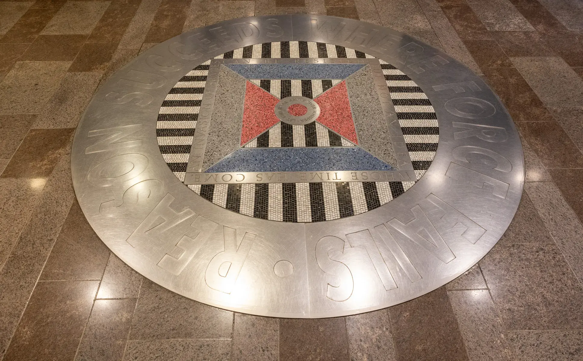 Circular floor installation with thick metal band around the edge and a black and white and red, blue, and gray mosaic and terrazzo pattern in the center. The words "Reason succeeds where force fails" are visible around the perimeter.