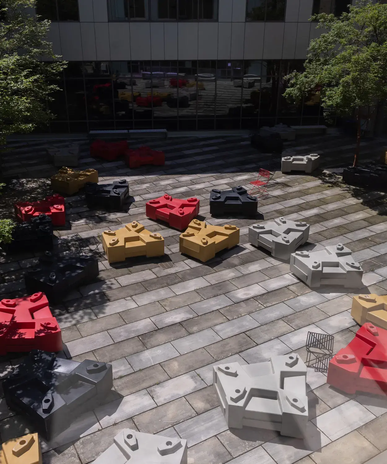 Red, yellow gold, gray, and black five-sided objects sit on a patio of light and dark gray pavers in alternating lines. There are trees on both sides.