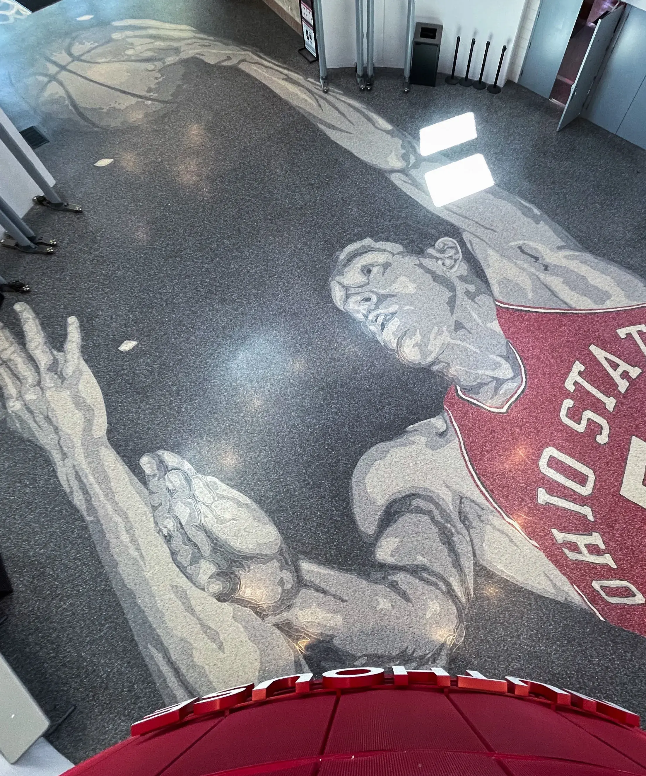 Floor design in grayscale and red shows a male basketball player wearing a red Ohio State 5 jersey making a layup. It is in a round space.