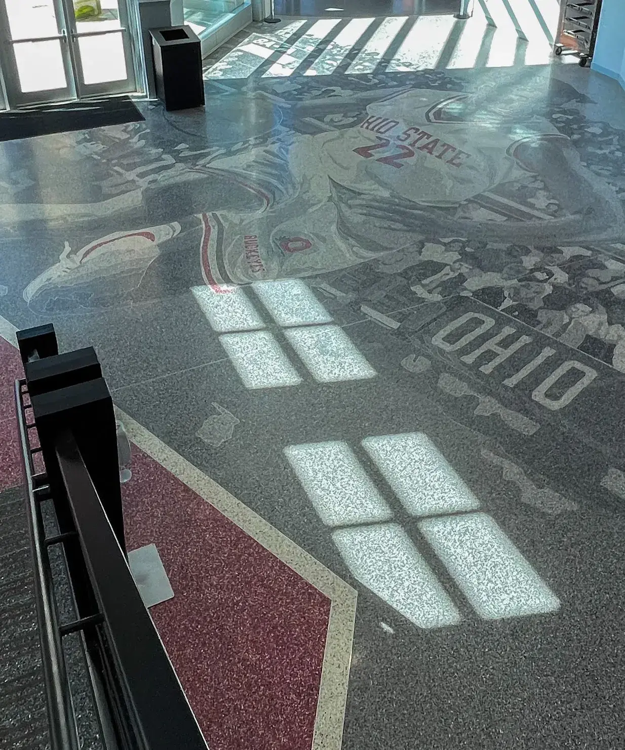 Floor design in grayscale and red shows a  basketball player wearing a white and red Ohio State 22 jersey dribbling the ball in front of crowded stands.