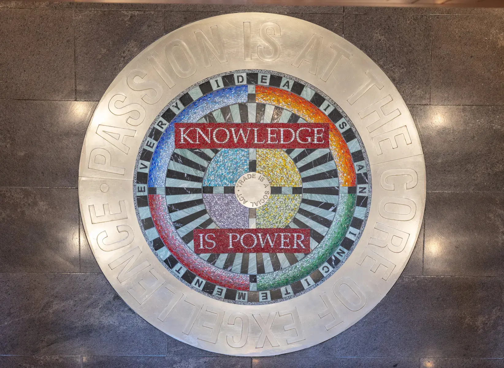 Circular floor installation with thick metal band around the edge and a black and white and multicolored mosaic pattern in the center. The words "knowledge is power" are visible.