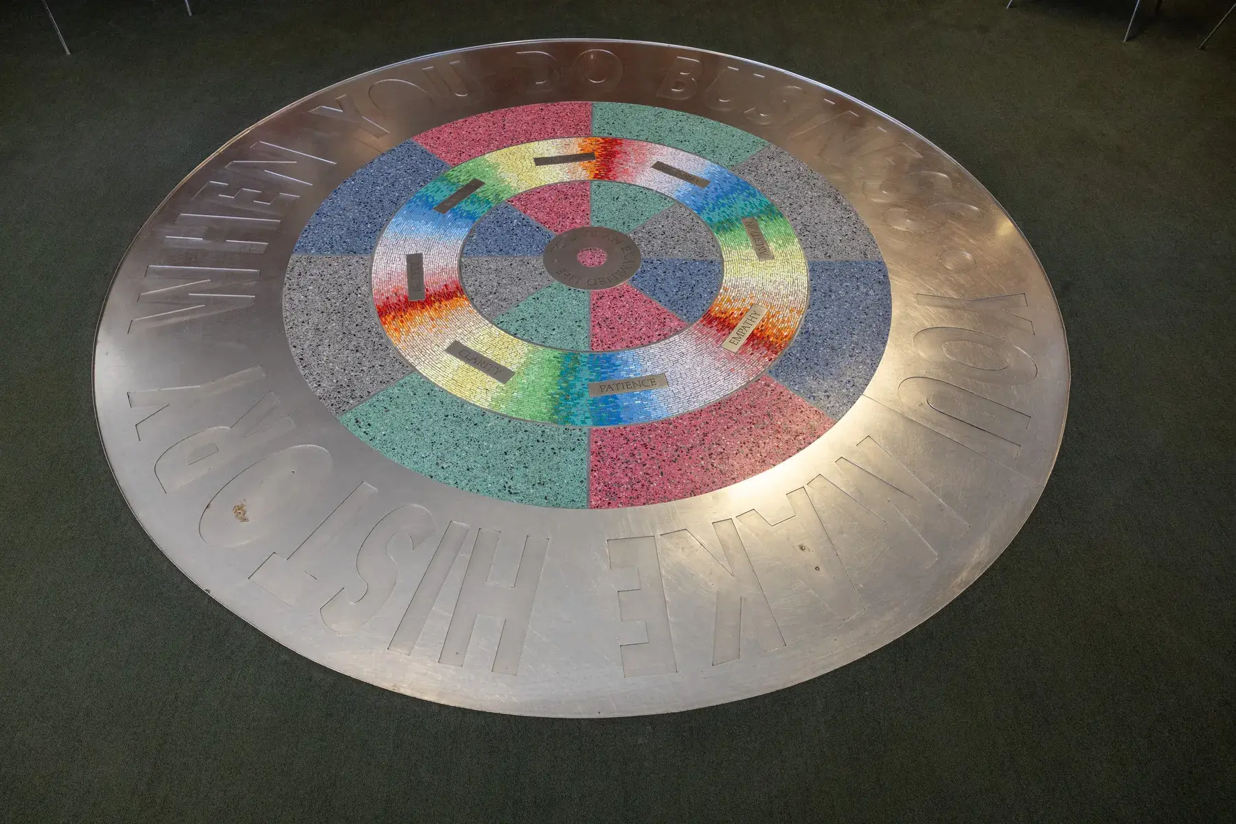 Circular floor installation with thick metal band around the edge and a multicolored mosaic and terrazzo pattern in the center. The words "You make history when you do business" are visible around the perimeter