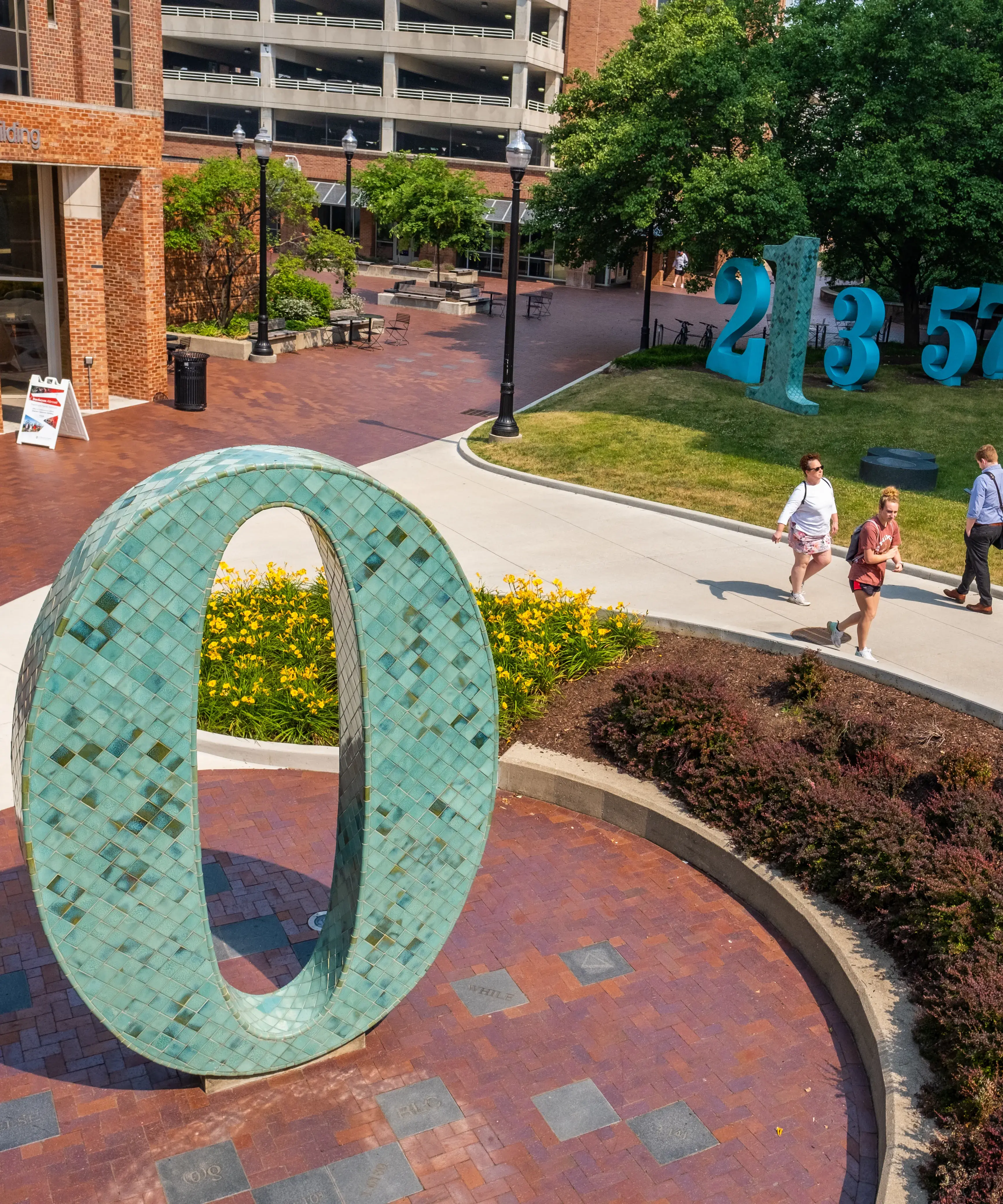 Group of large sculptural numbers in blue, gold, and tiles of green on both sides of a walkway. The zero in the foreground sits in a brick courtyard with small metal plaques embedded on the ground.