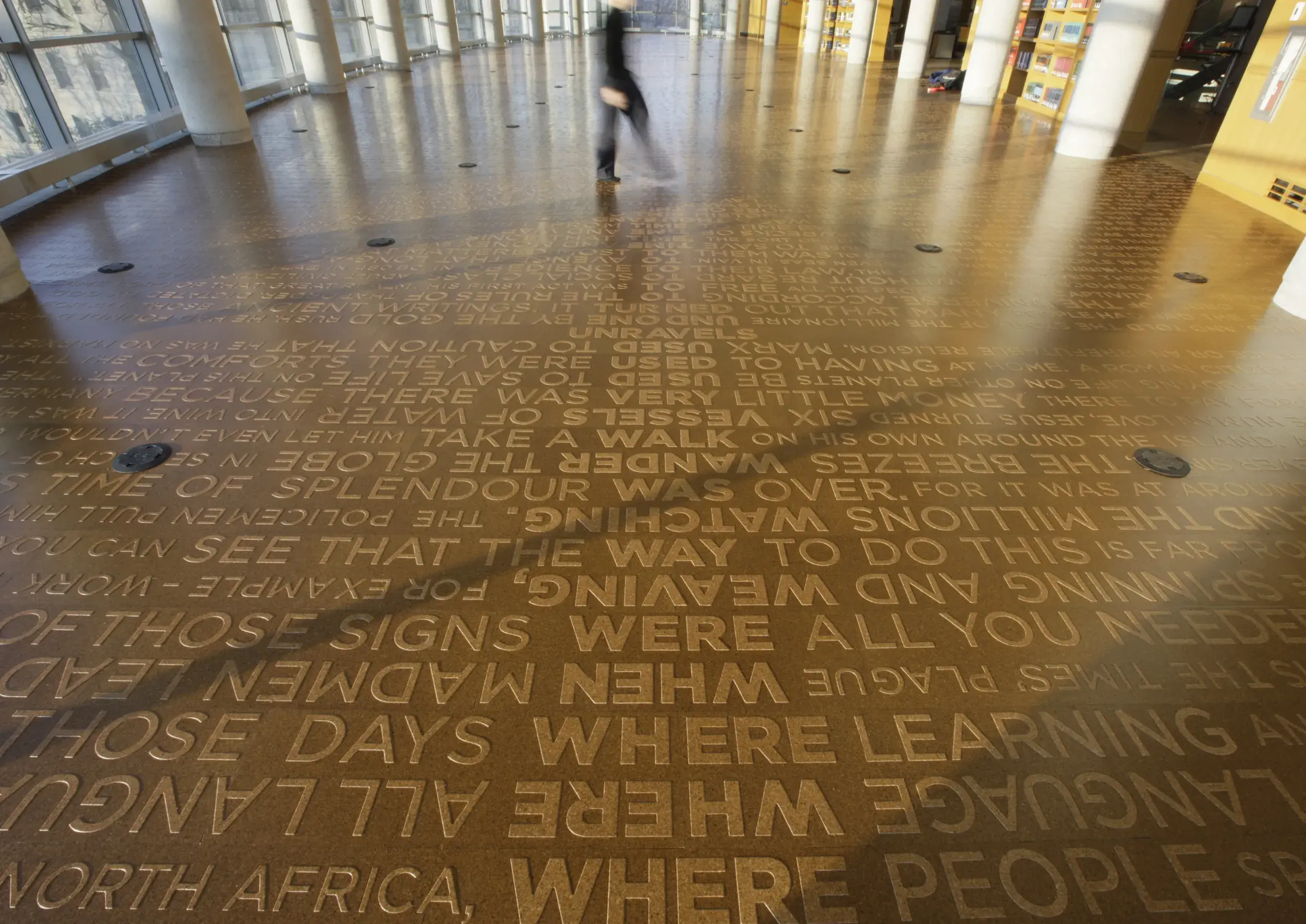 Medium brown background with rows of lighter brown words slightly raised up from the surface displayed on the floor in a large open room