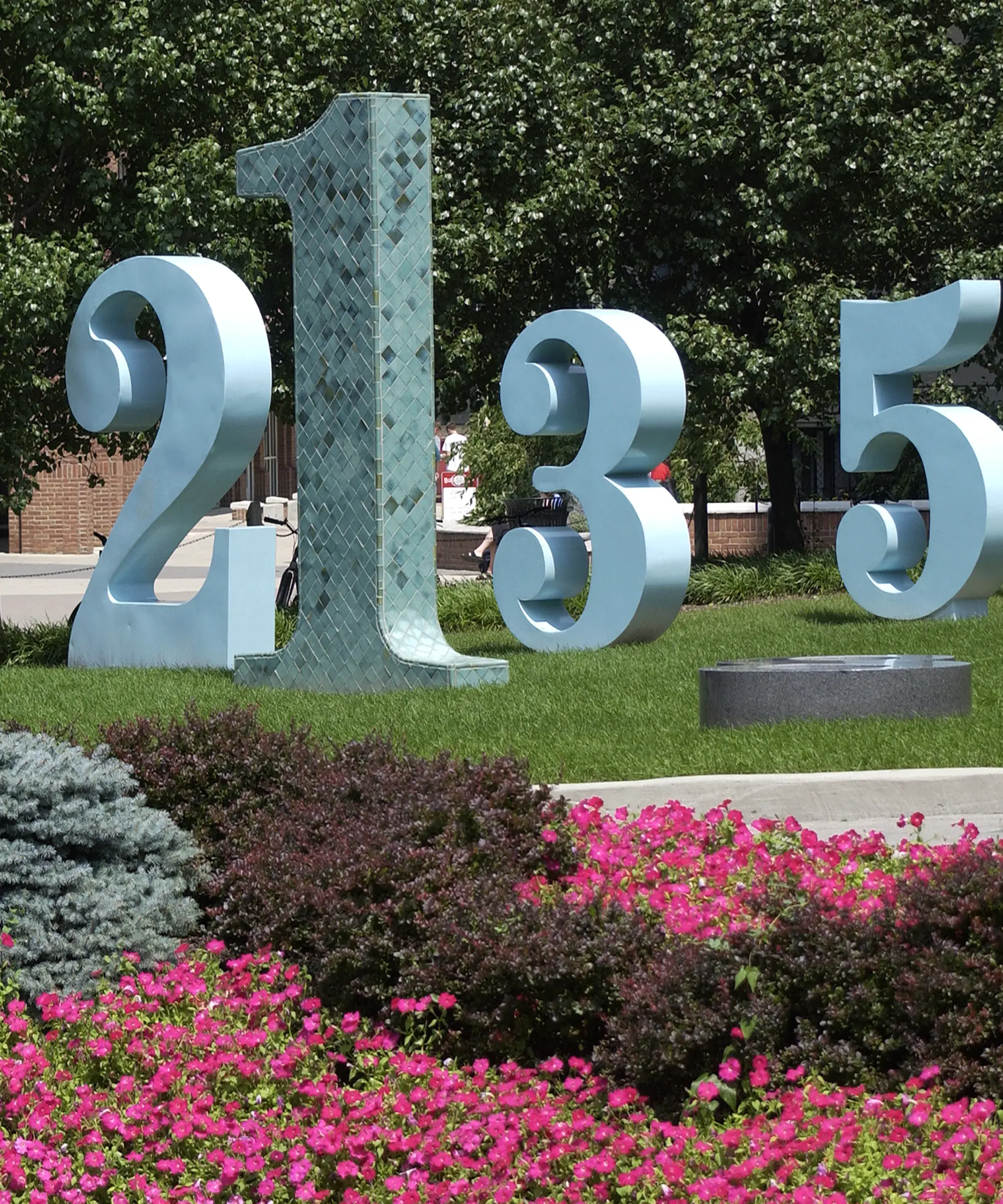 A group of large sculptural numbers in blue and tiles of green sit in a grassy area with flowers and bushes in the foreground