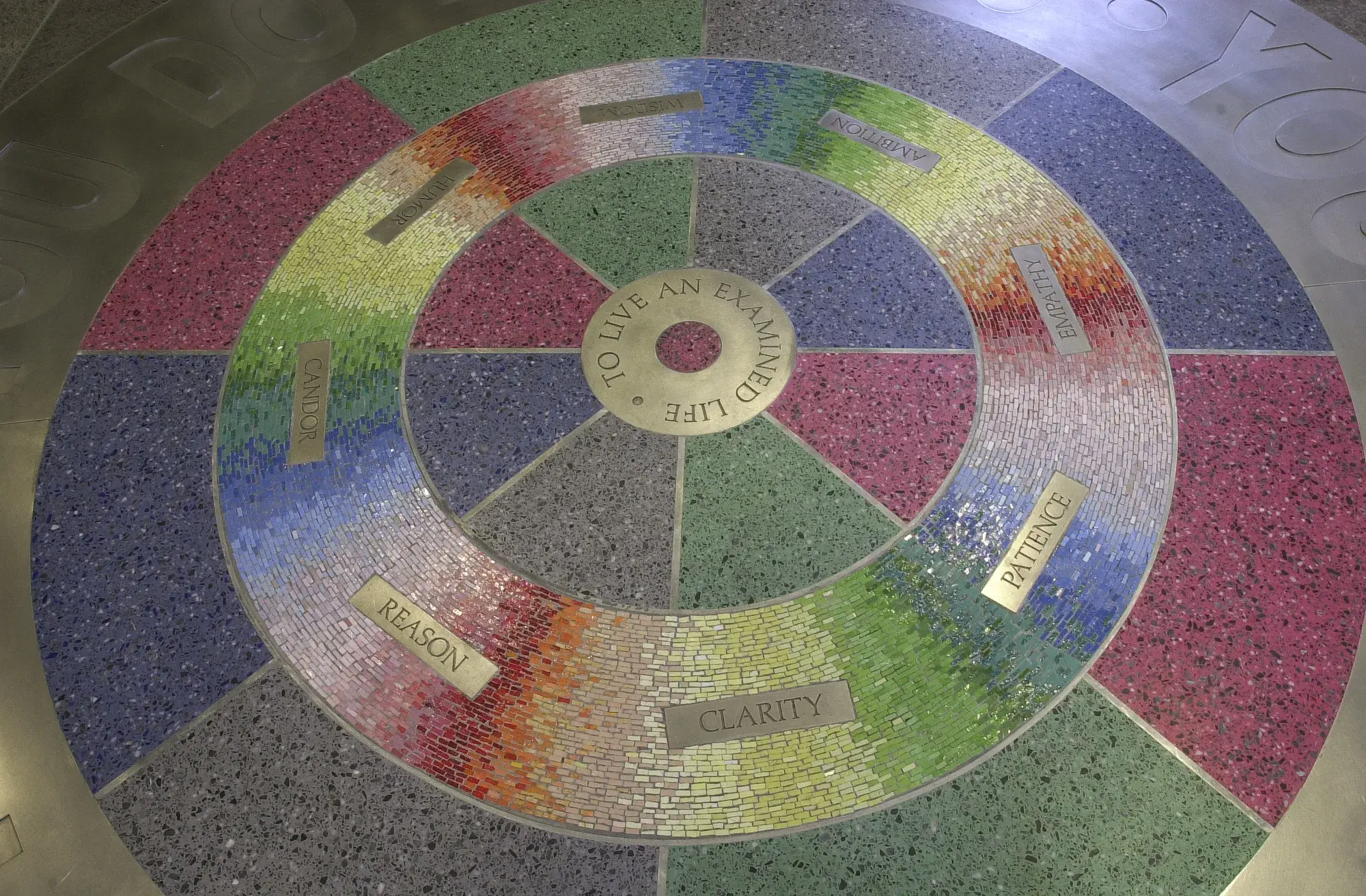 Circular floor installation with thick metal band around the edge and a multicolored mosaic and terrazzo pattern in the center. The words "To Live an Examined Life" are visible in the center.