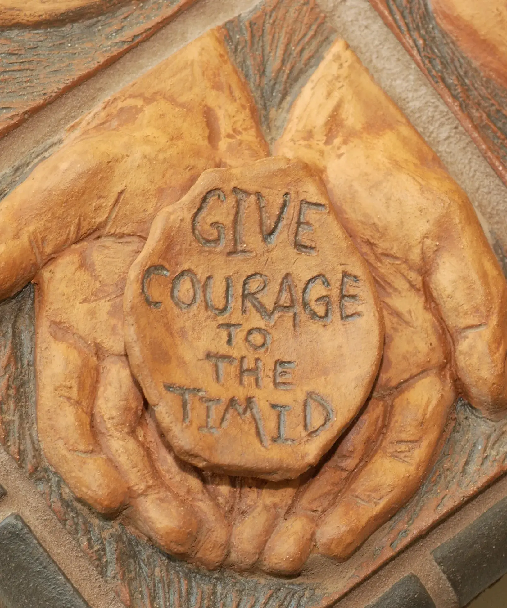 A closeup of a tile on the corner of an artwork which shows two hands holding an oval that says "Give Courage to the Timid."