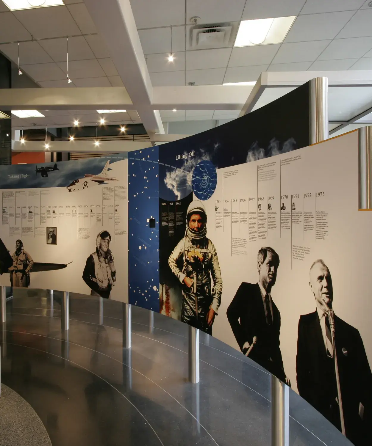 Images of planes, astronauts and men in suits and a timeline on a white background are printed on a curved panels held up on metal legs. Another panel has an image of metal horn or bell on it. The shiny floor has rings on it.