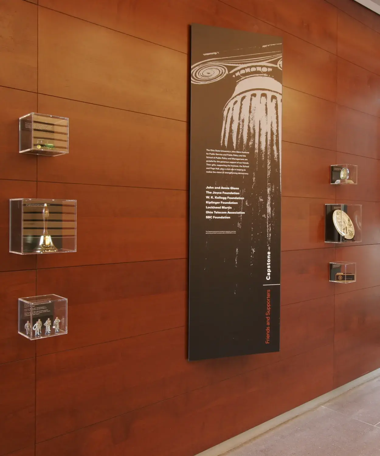 Small clear plastic cases with objects in them and larger black and white panels are displayed on a wood-covered wall.