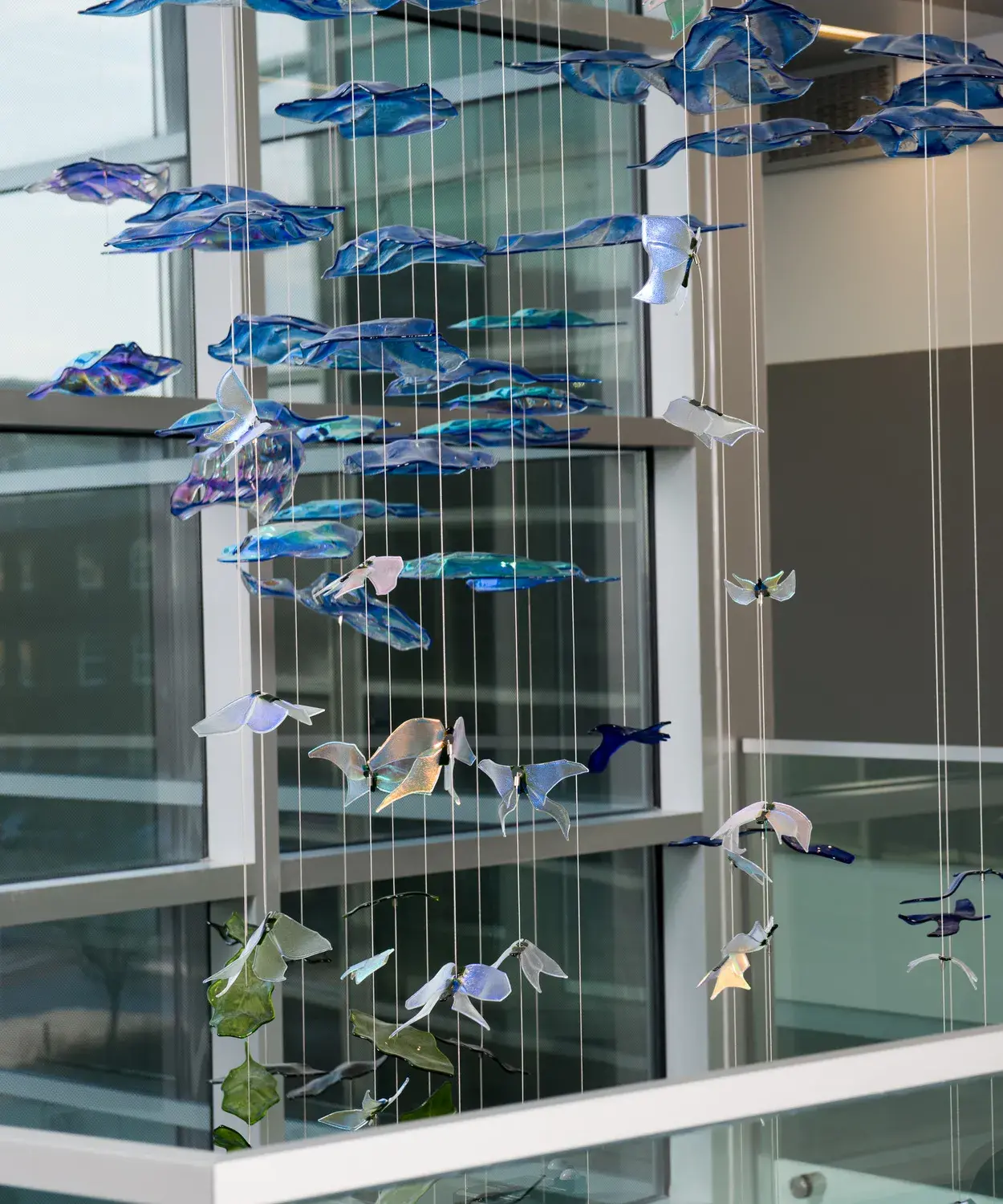 Small wavy glass pieces in blue, white, and green in abstract shapes reminiscent of birds and butterflies are suspended on white cables through a glass-framed atrium space bordered by a glass railing.