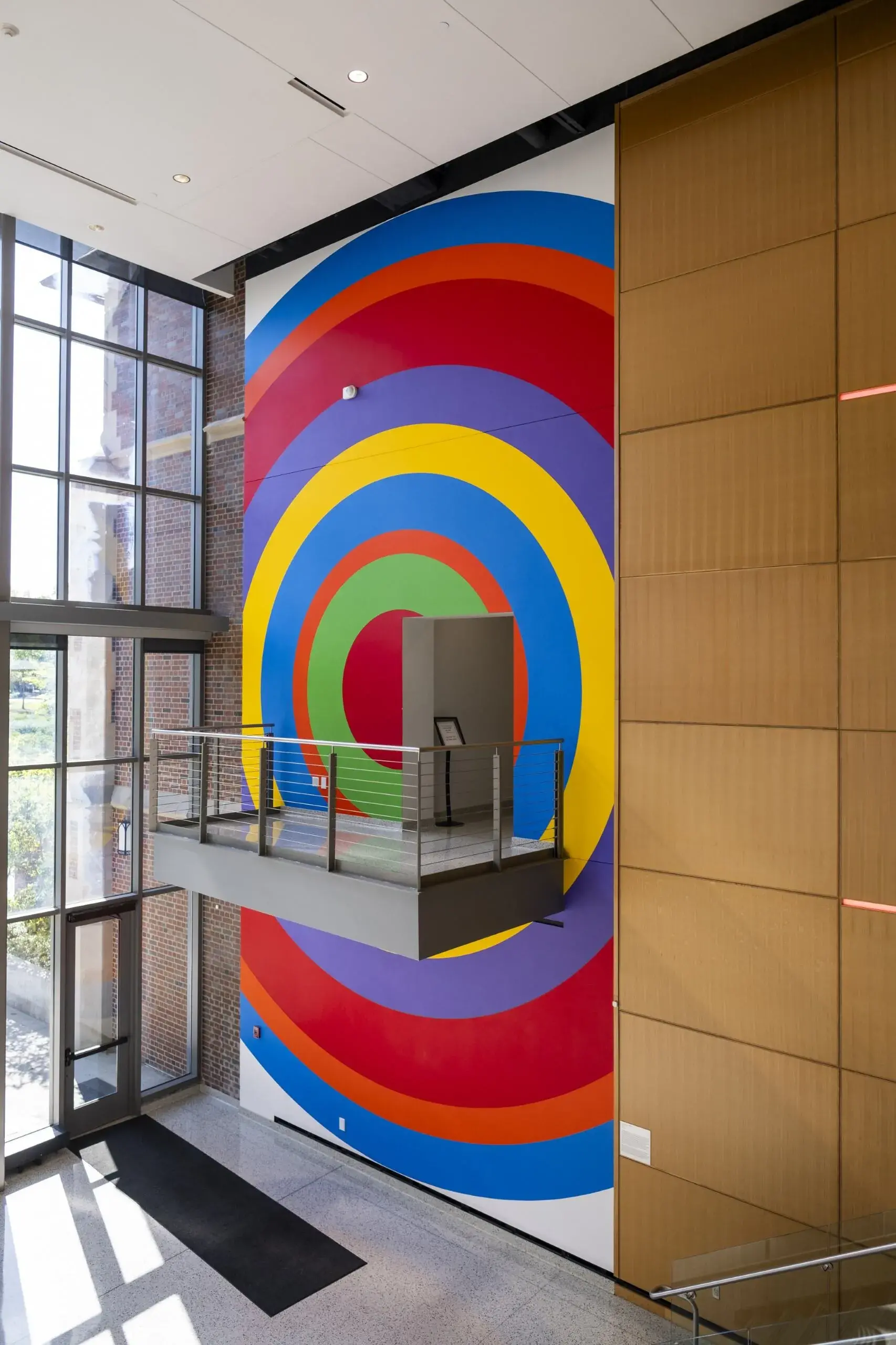 Multistory atrium space with full height wall painting in one section near the windows. The painting is made of concentric circles of vibrant colors. There is a small balcony near the middle of the painting.