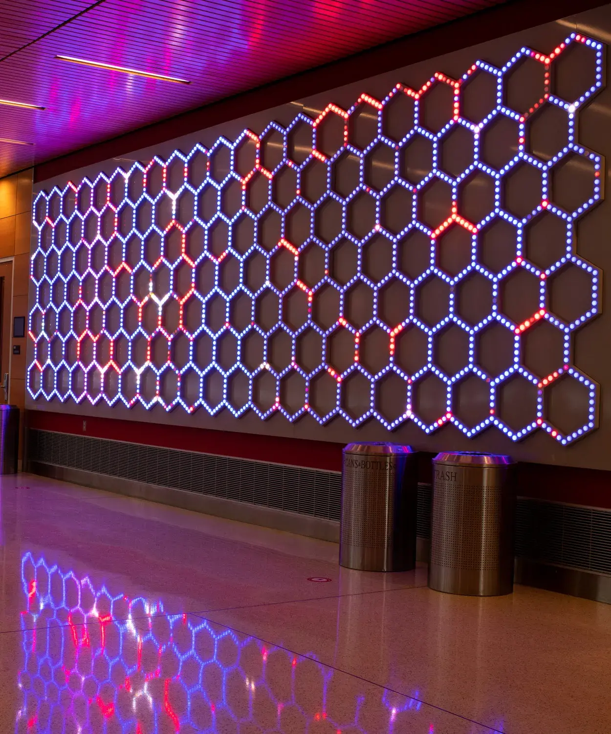 Lobby space with three walls of a light installation consisting of small bulbs in a honeycomb pattern. The lights have different patterns of red and bluish purple and reflect onto the floor and ceiling.