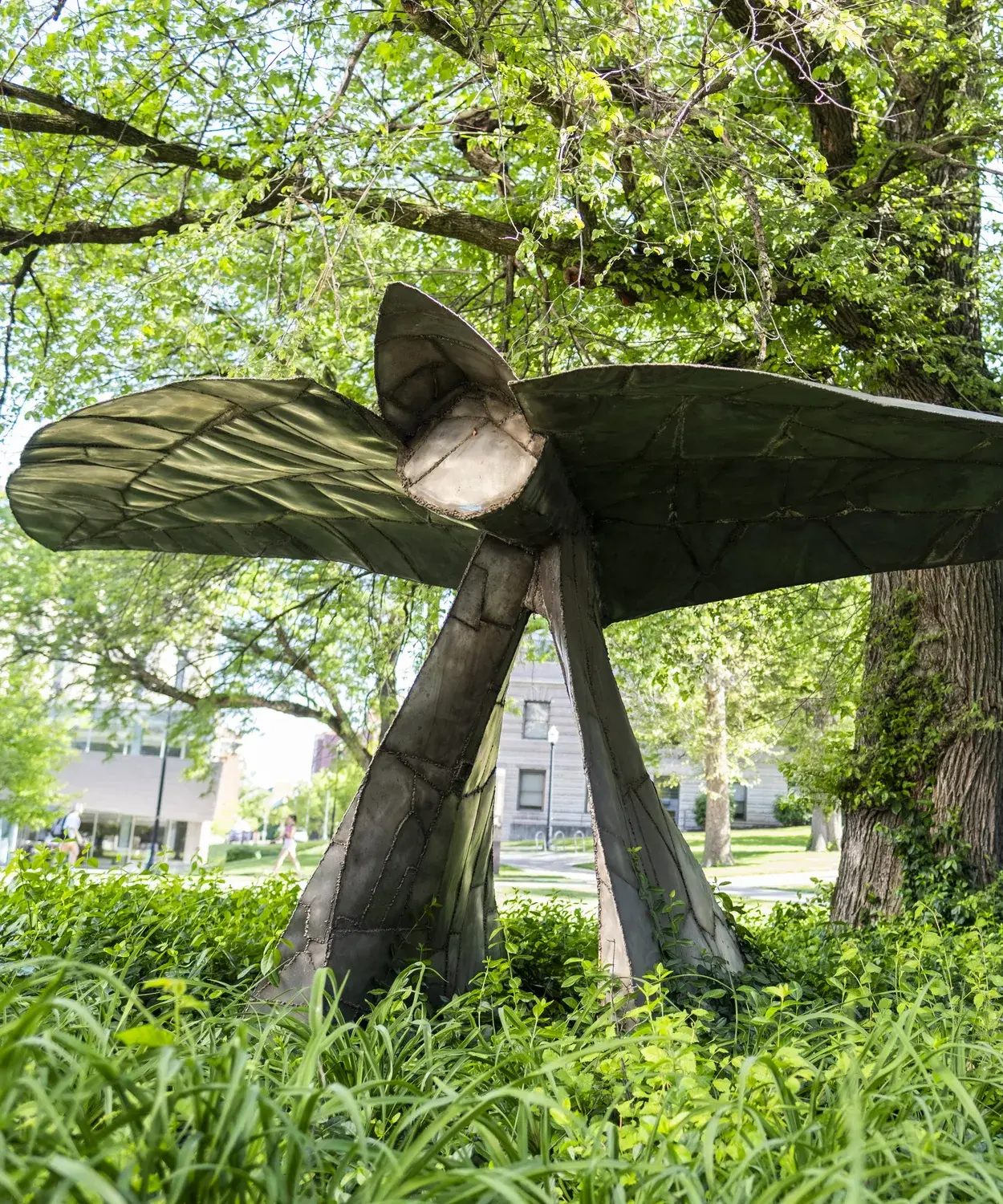 Metal sculpture sits in a landscape bed under a tree. The sculpture has a horizontal component on top of two triangular vertical base pieces and a circular part near the middle.