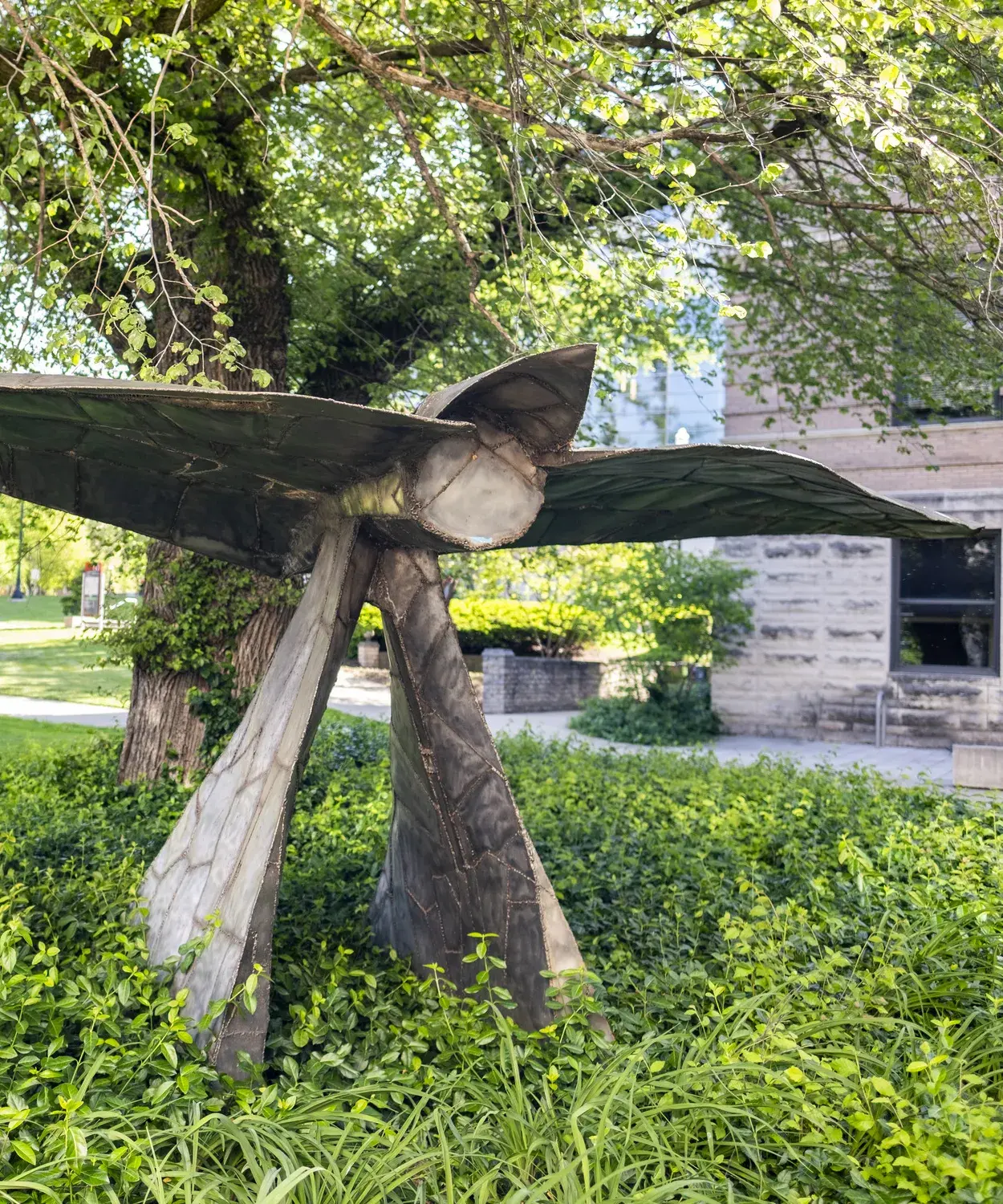 Metal sculpture sits in a landscape bed under a tree. The sculpture has a horizontal component on top of two triangular vertical base pieces and a circular part near the middle.