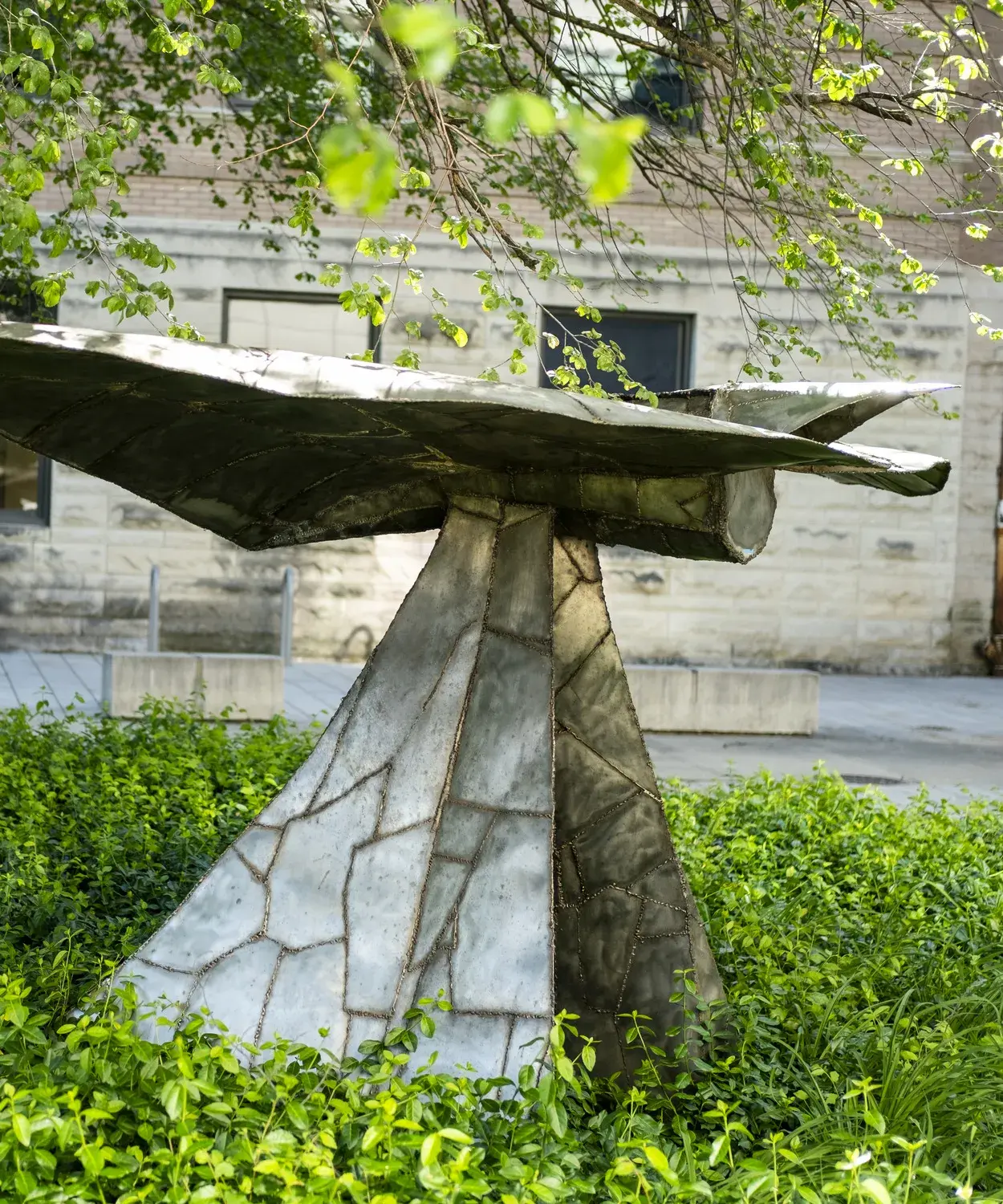 Metal sculpture sits in a landscape bed next to a tree near a stone building. The sculpture has a horizontal component on top of a triangular vertical base.