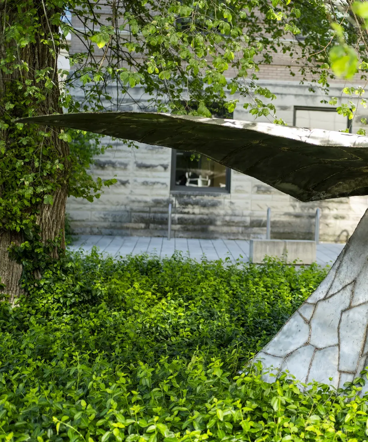 Metal sculpture sits in a landscape bed next to a tree near a stone building. The sculpture has a horizontal component on top of a triangular vertical base.