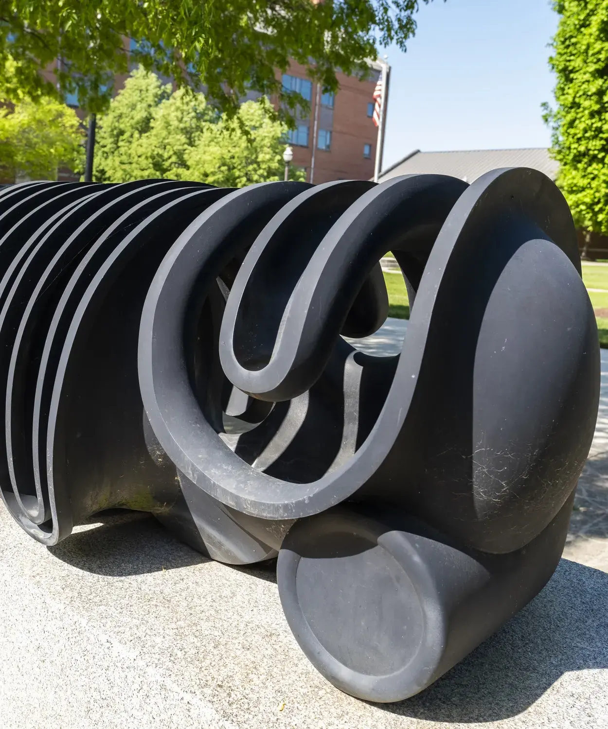 Dark metal sculpture of curvilinear forms sits on a gray stone base with leafy green trees and a building behind it