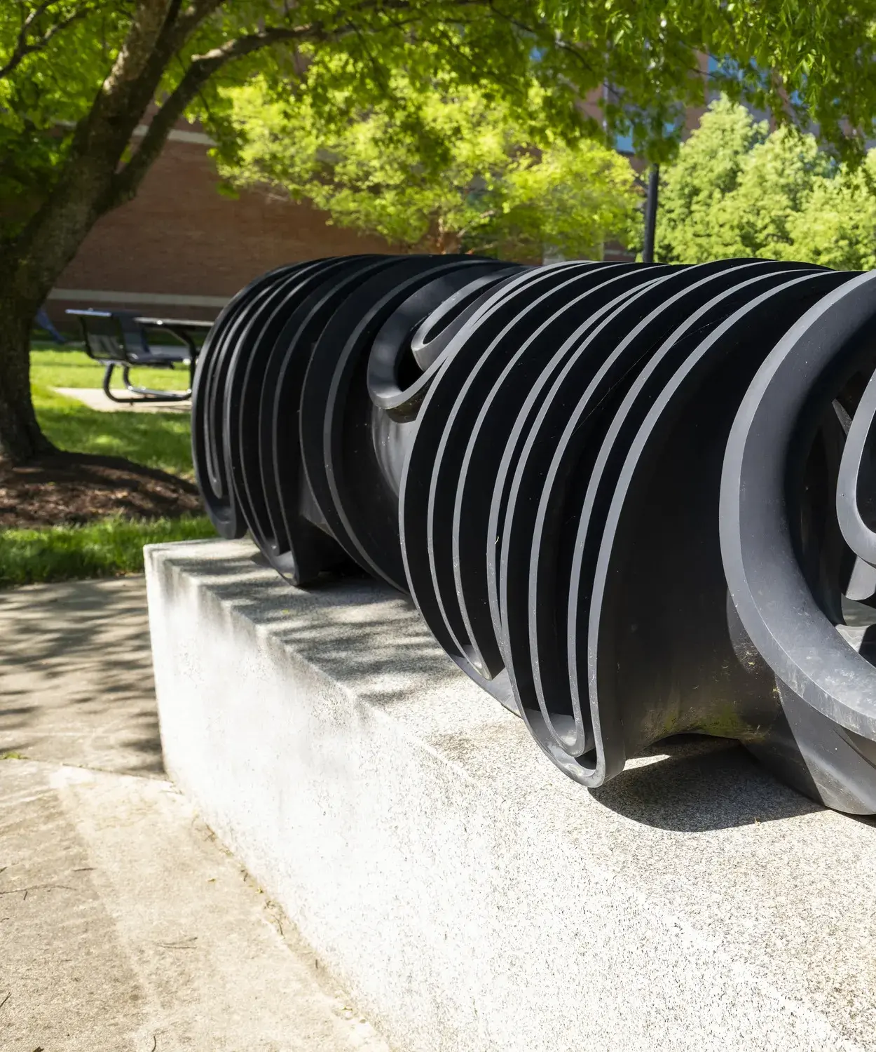Dark metal sculpture of curvilinear forms sits on a gray stone base with leafy green trees and a building behind it
