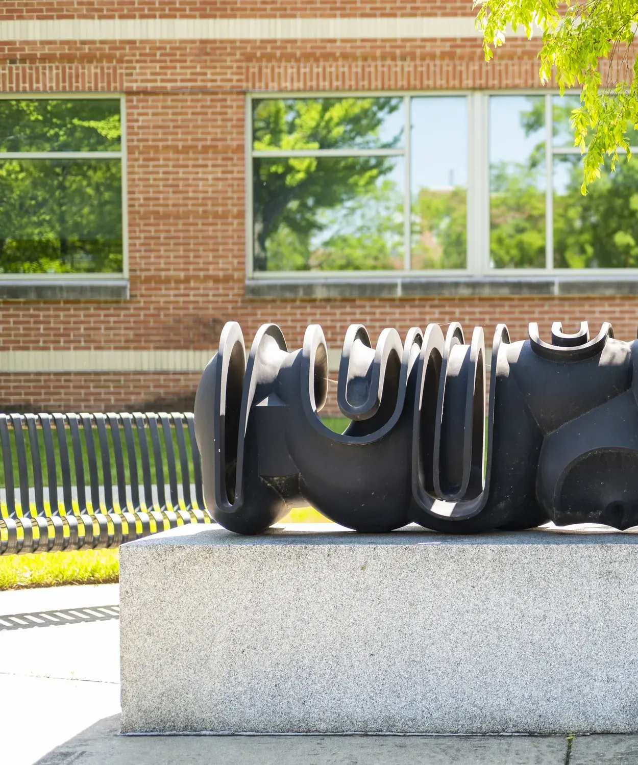 Dark metal sculpture of curvilinear forms sits on a gray stone base with leafy green trees and a building behind it