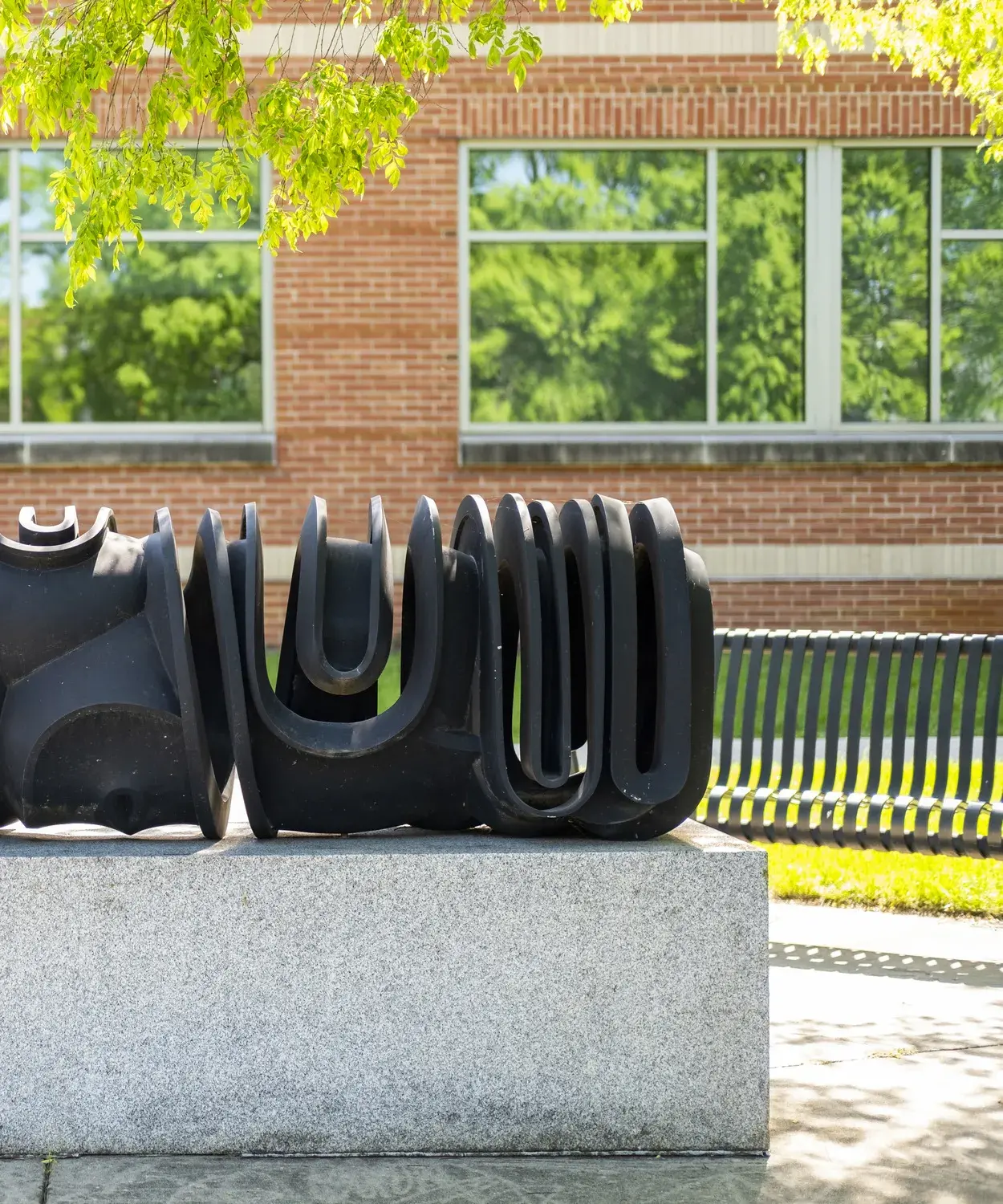 Dark metal sculpture of curvilinear forms sits on a gray stone base with leafy green trees and a building behind it