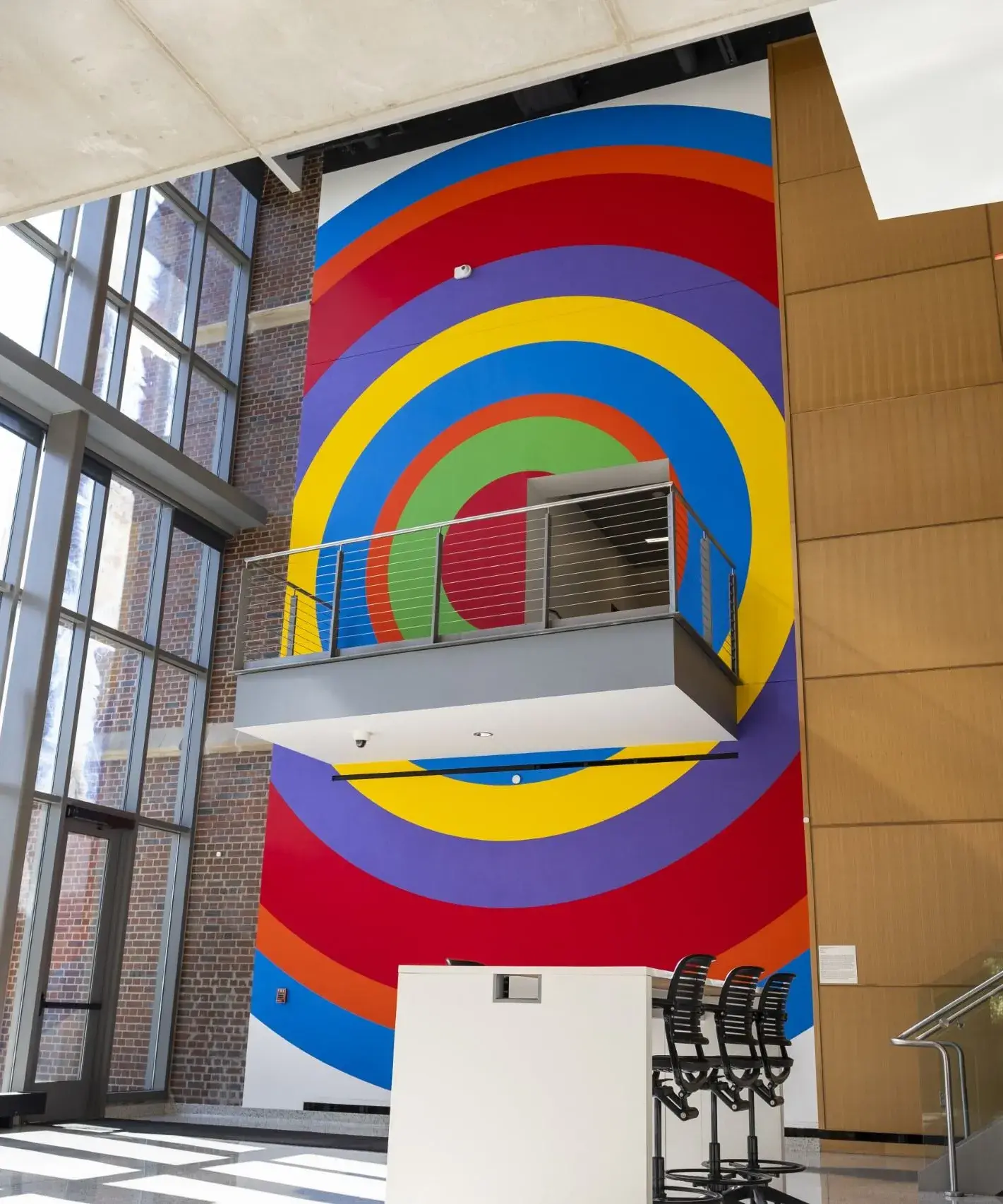 Multistory atrium space with full height wall painting in one section near the windows. The painting is made of concentric circles of vibrant colors. There is a small balcony near the middle of the painting.