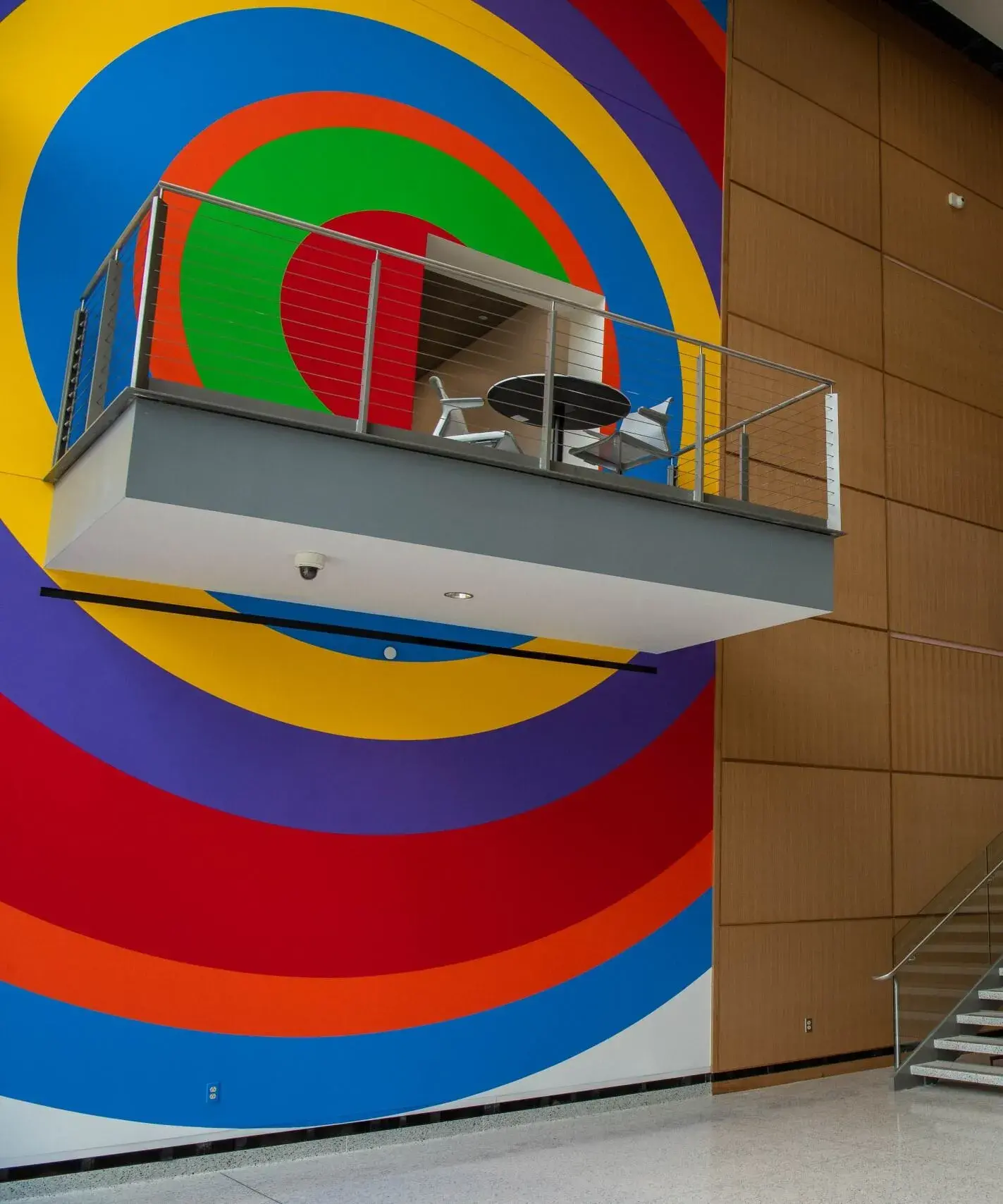 Multistory atrium space with full height wall painting in one section near the windows. The painting is made of concentric circles of vibrant colors. There is a small balcony near the middle of the painting and there are other balconies and stairs in the image.