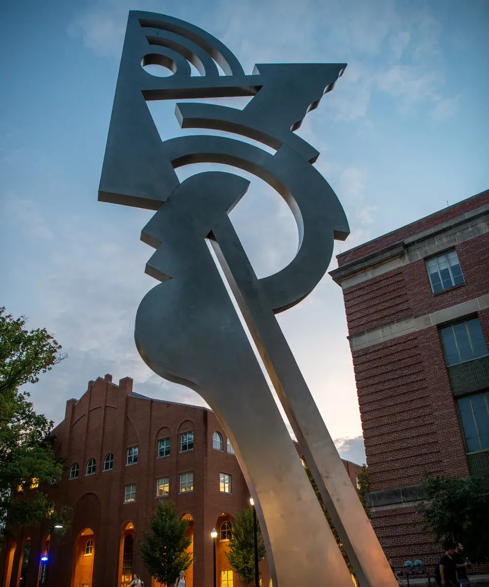 Large silver colored sculpture in the shape of an abstract head on a long neck. There are several-story-high brick buildings and a dusk sky in the background. The photo is taken from ground level, looking up.