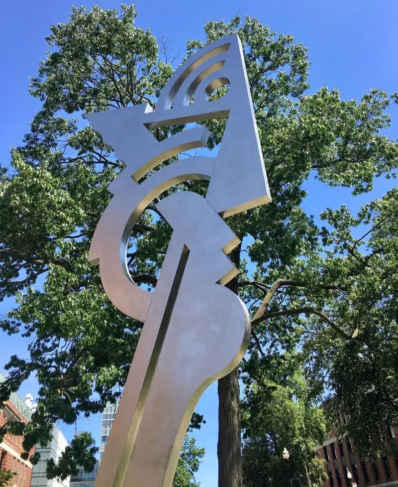 Large silver colored sculpture in the shape of an abstract head on a long neck. There are several-story-high brick buildings and a dusk sky in the background. The photo is taken from ground level, looking up.