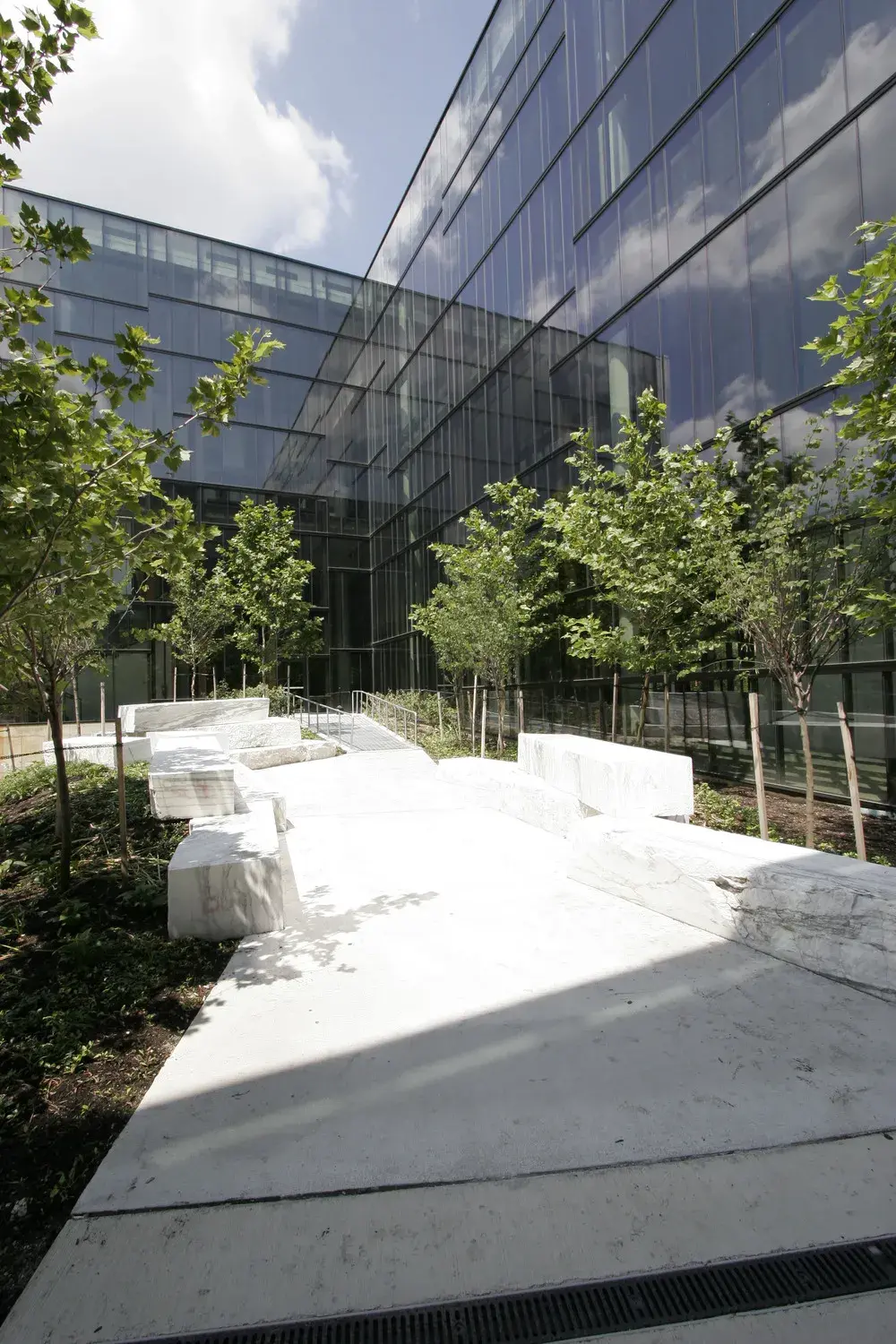Slabs of white and gray marble which sit alongside a walkway in a courtyard with trees, next to a glass building wall, with bright sun shining in