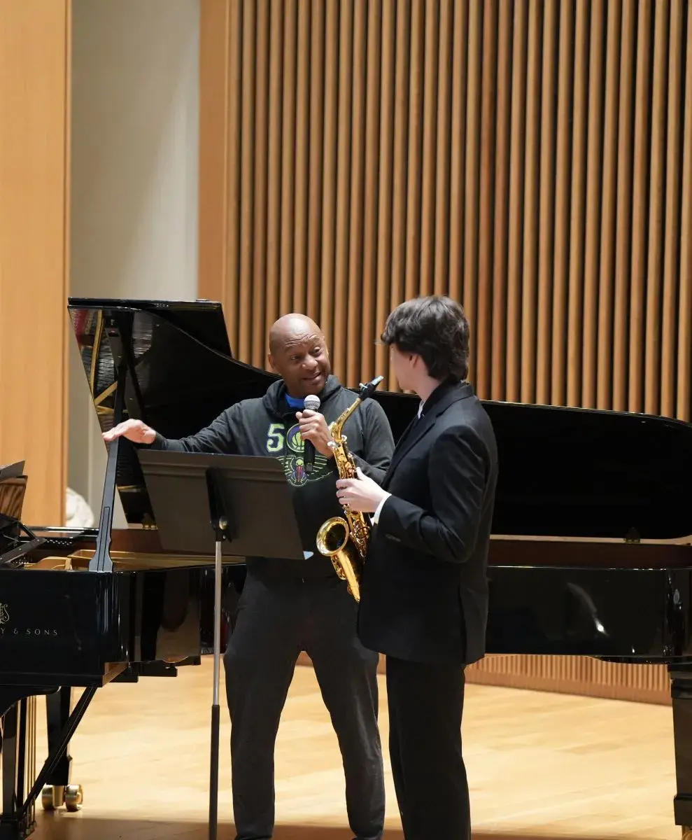 Brandford Marsalis speaks with a student holding a saxophone in front of a piano.