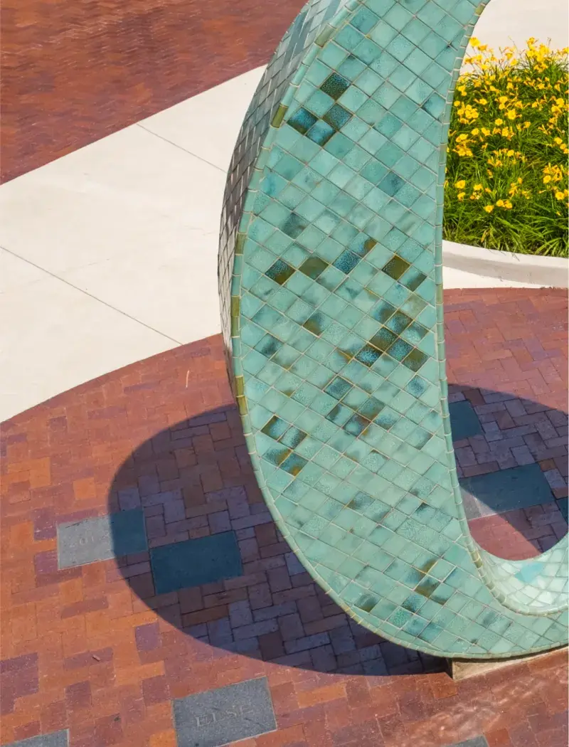 A close up of a sculptural number zero in blue, gold, and tiles of green and positioned in a brick courtyard with small metal plaques embedded on the ground.