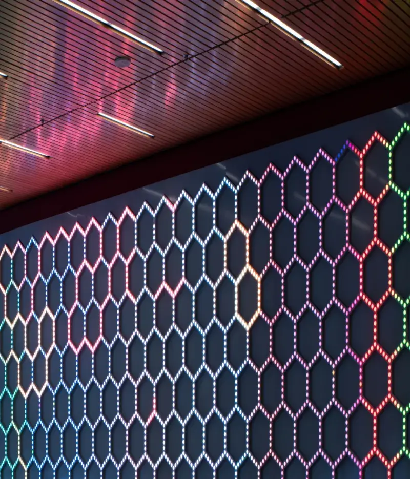 Detailed image of a lobby space with three walls of a light installation consisting of small bulbs in a honeycomb pattern. The lights have different patterns of red and bluish purple and reflect onto the floor and ceiling.